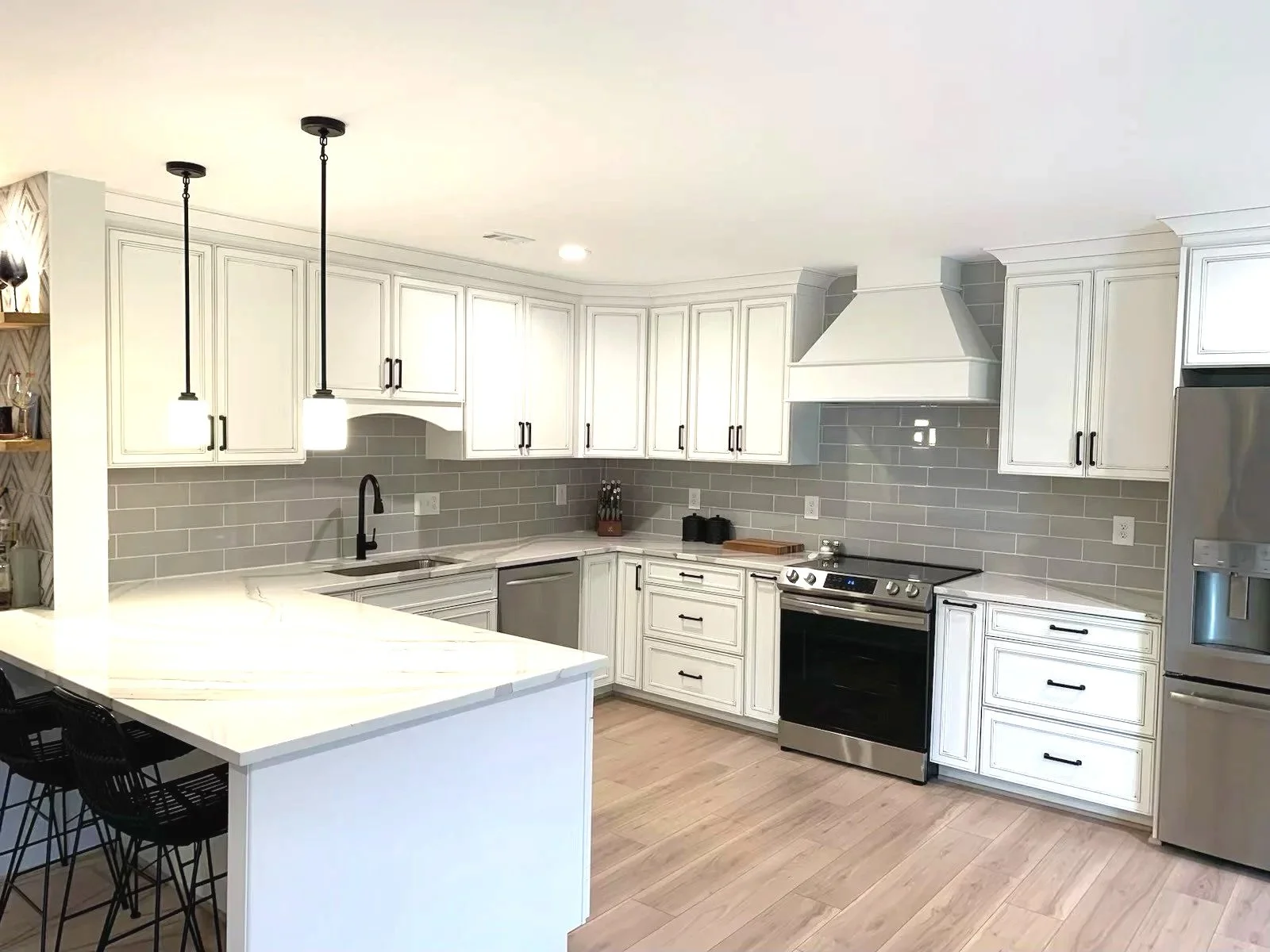 Modern white kitchen with grey subway tile backsplash, stainless steel appliances, black hardware, and pendant lighting over a white island with black bar stools.