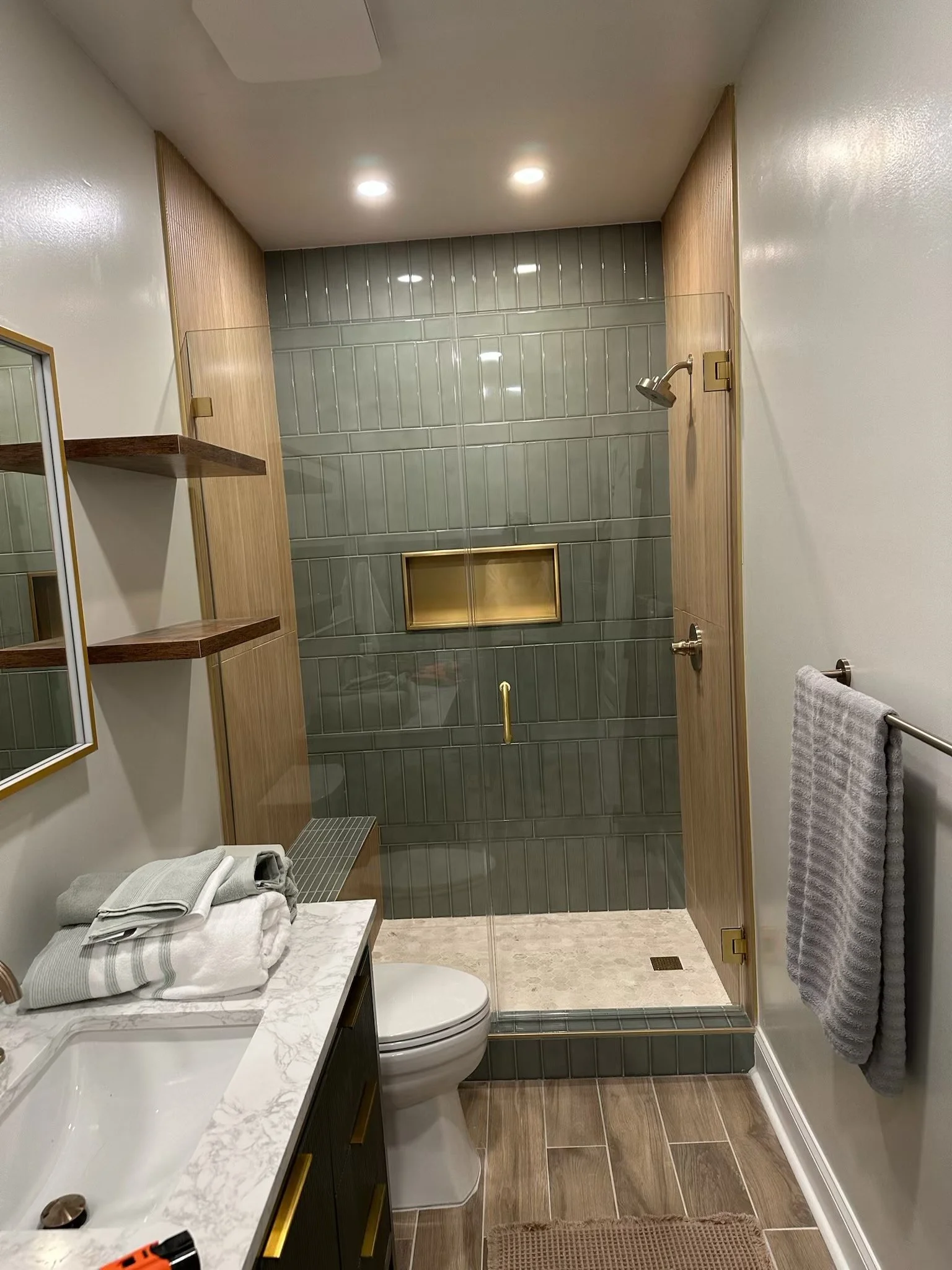 A modern bathroom with a glass-enclosed shower featuring gray tile and a built-in niche. The bathroom has a white countertop with a sink, folded towels, a mirror, and a towel rack with a gray towel. The floor has wood-look tiles, and the ceiling has recessed lighting.