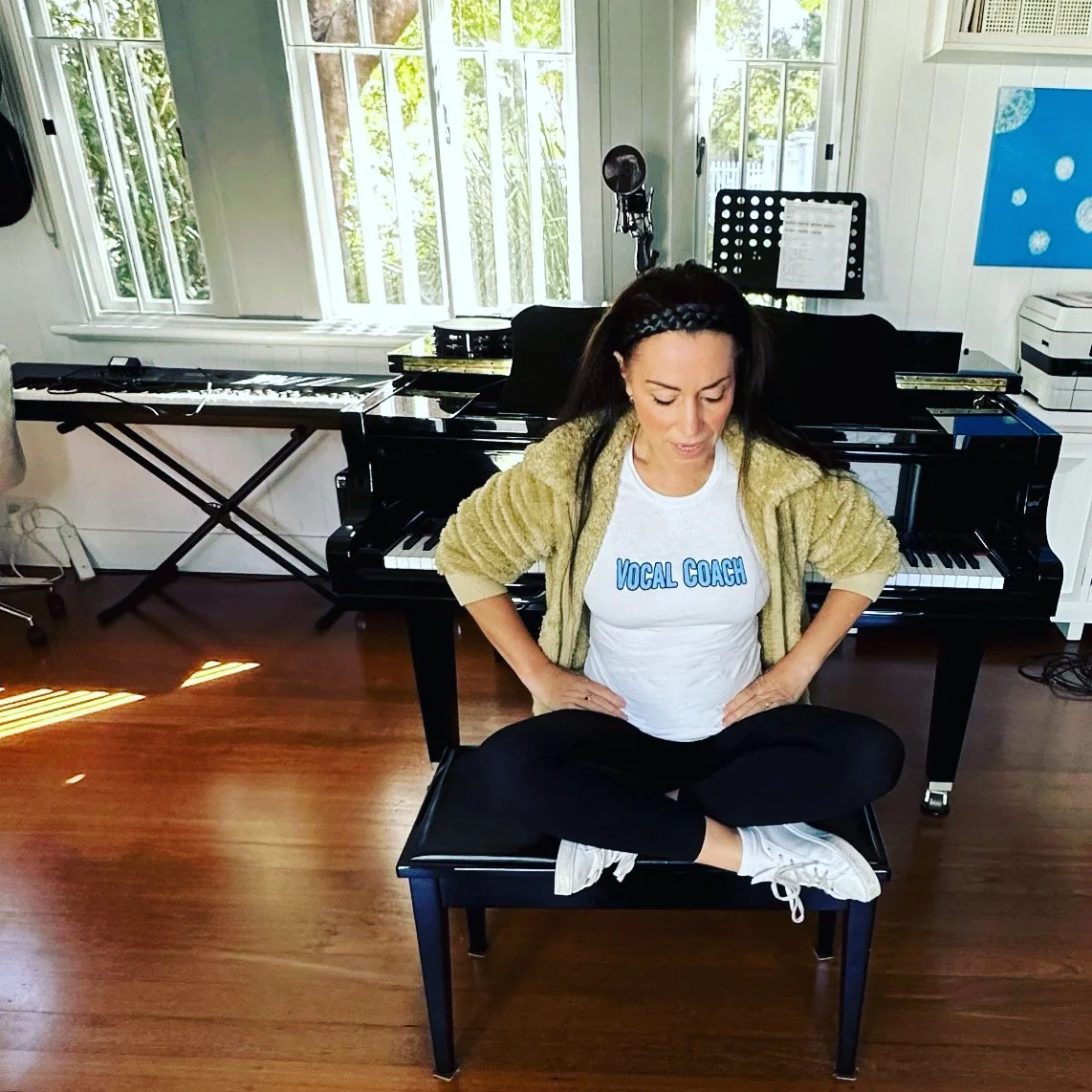 A woman sitting cross-legged on a piano bench in a music studio, with a grand piano and a keyboard in the background, wearing a white t-shirt that says 'Vocal Coach' and a beige cardigan.