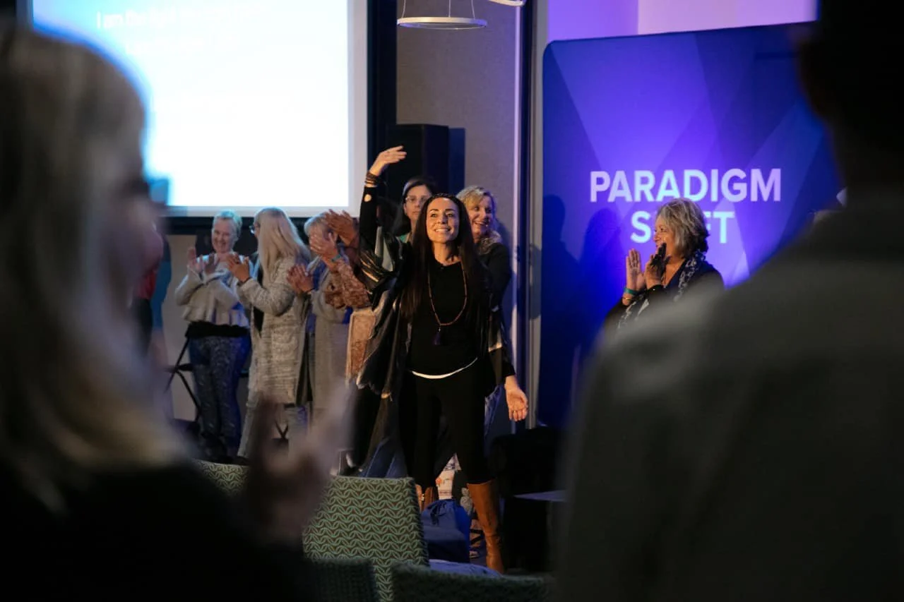 A group of women, mostly middle-aged, standing on a stage at a conference or event, clapping and smiling. One woman in the foreground has dark hair, is dressed in black, and is waving or gesturing. Behind her, a purple banner reads 'PARADIGM SHIFT.' The setting appears to be a professional gathering or seminar.