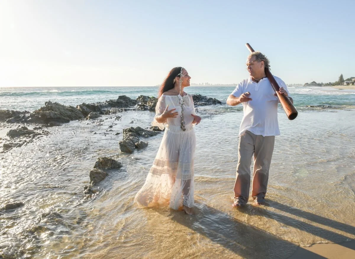 A woman in a white dress and a man with a woodwind instrument stand on a beach, engaging in conversation. The woman is smiling, and the man has a casual posture. The ocean, rocks, and a clear sky are in the background.