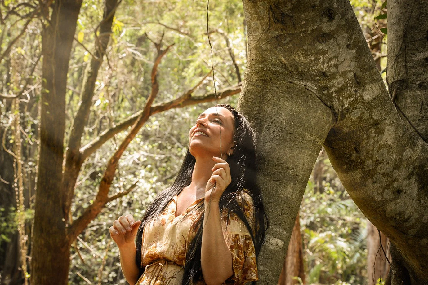 A woman with long dark hair smiling and looking up while standing outdoors in a forest, holding a thin string or vine near her face.