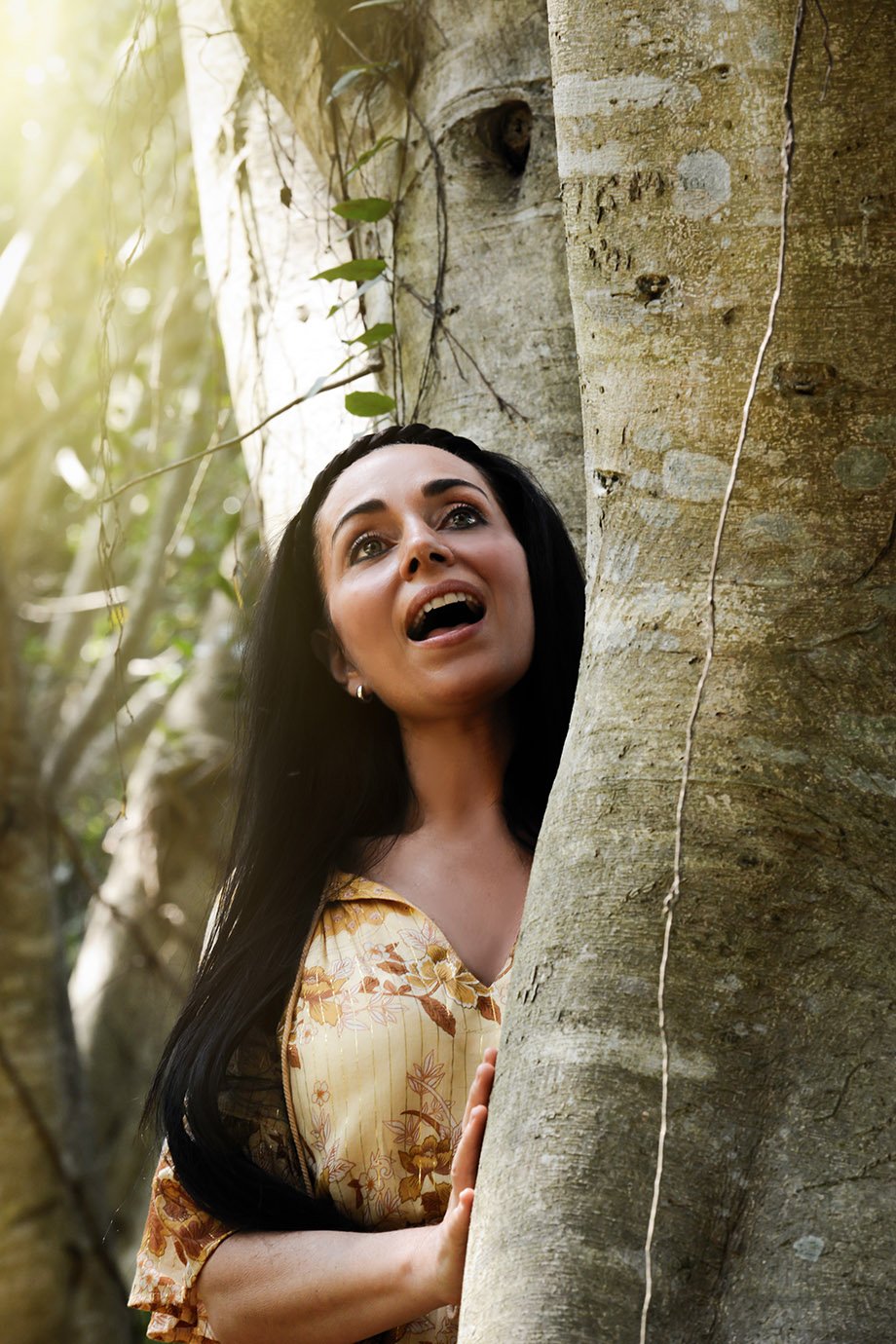 A woman with long black hair and a floral top hiding behind a tree, looking surprised or curious in a wooded area.