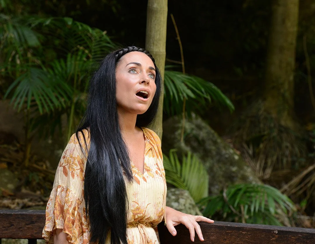 Woman with long black hair, wearing a yellow floral dress, standing outdoors next to a wooden railing in a forested area.