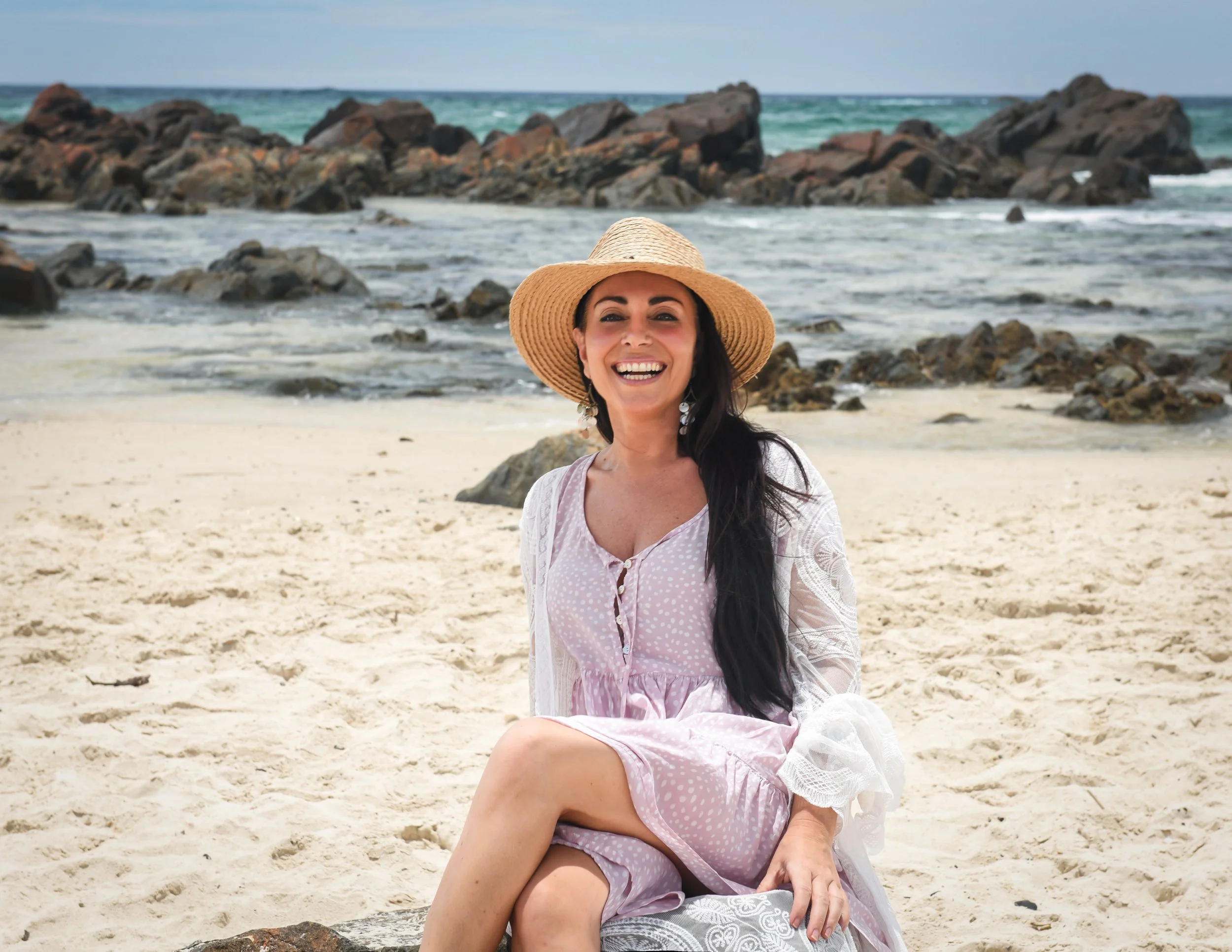 Woman in pink dress sitting on sandy beach with rocks and ocean in background, wearing a wide-brimmed straw hat and smiling