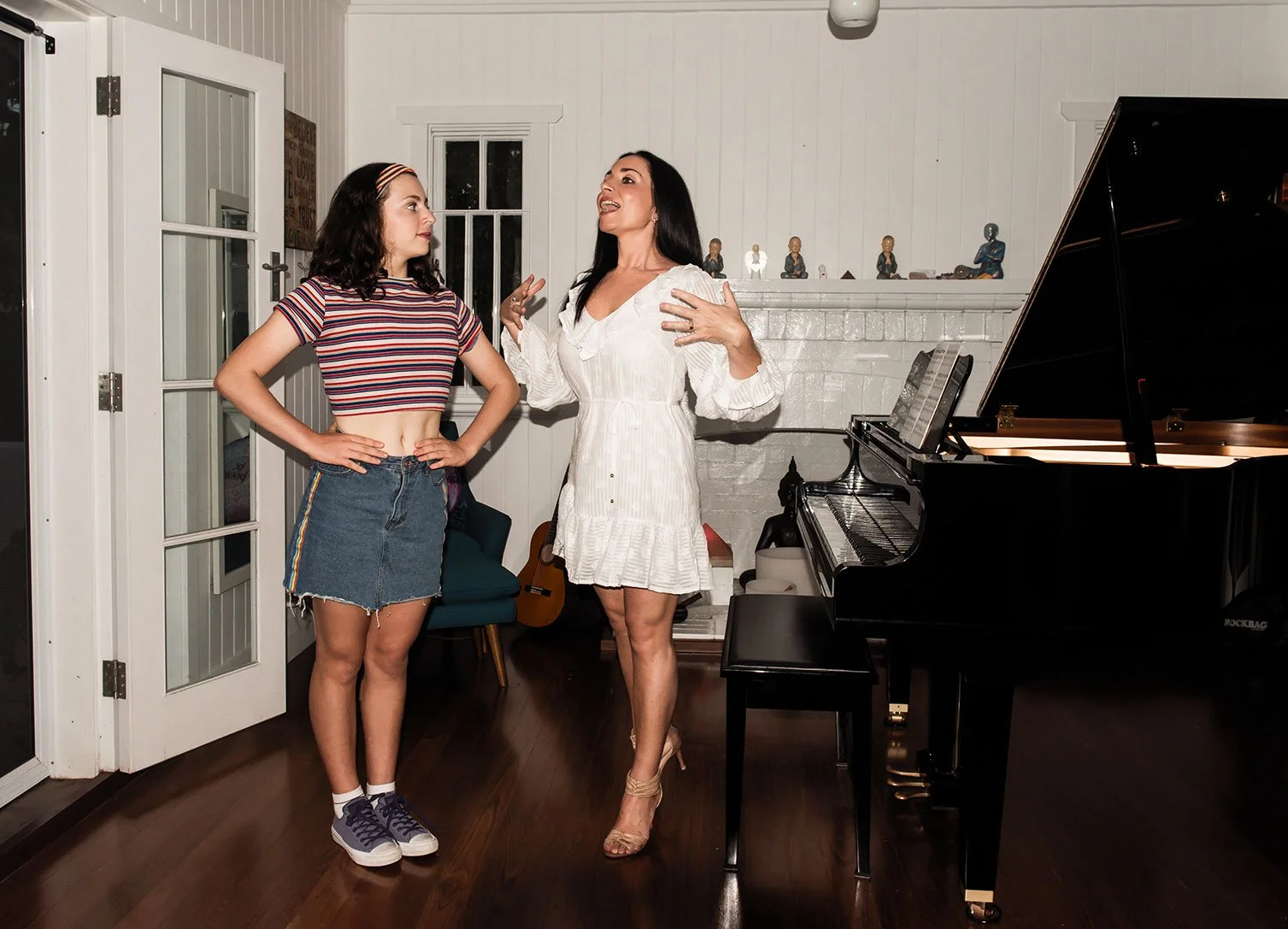 Two women having a conversation in a room with a black grand piano, a window, and a brick fireplace. One woman is young, wearing a striped crop top and denim skirt, and the other woman is older, wearing a white dress, gesturing as she speaks.