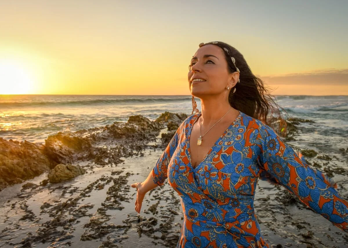 A woman at the beach during sunset, wearing a blue and orange floral dress with her arms outstretched and a serene expression.