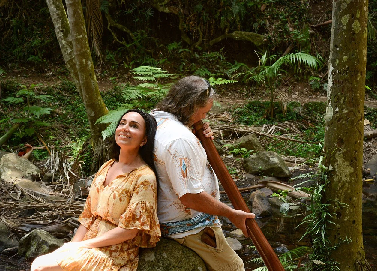 A man with long hair playing a wooden flute while sitting on a rock in a lush, green forest next to a woman with long dark hair in a yellow floral dress, who is smiling and looking up.