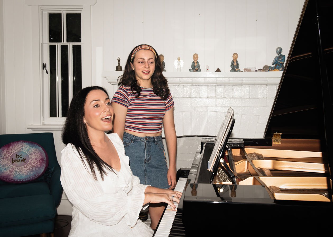 Two women, one playing a grand piano and the other singing, in a cozy living room with decorative figures on a mantle and a pillow that says 'be brave' on a sofa.