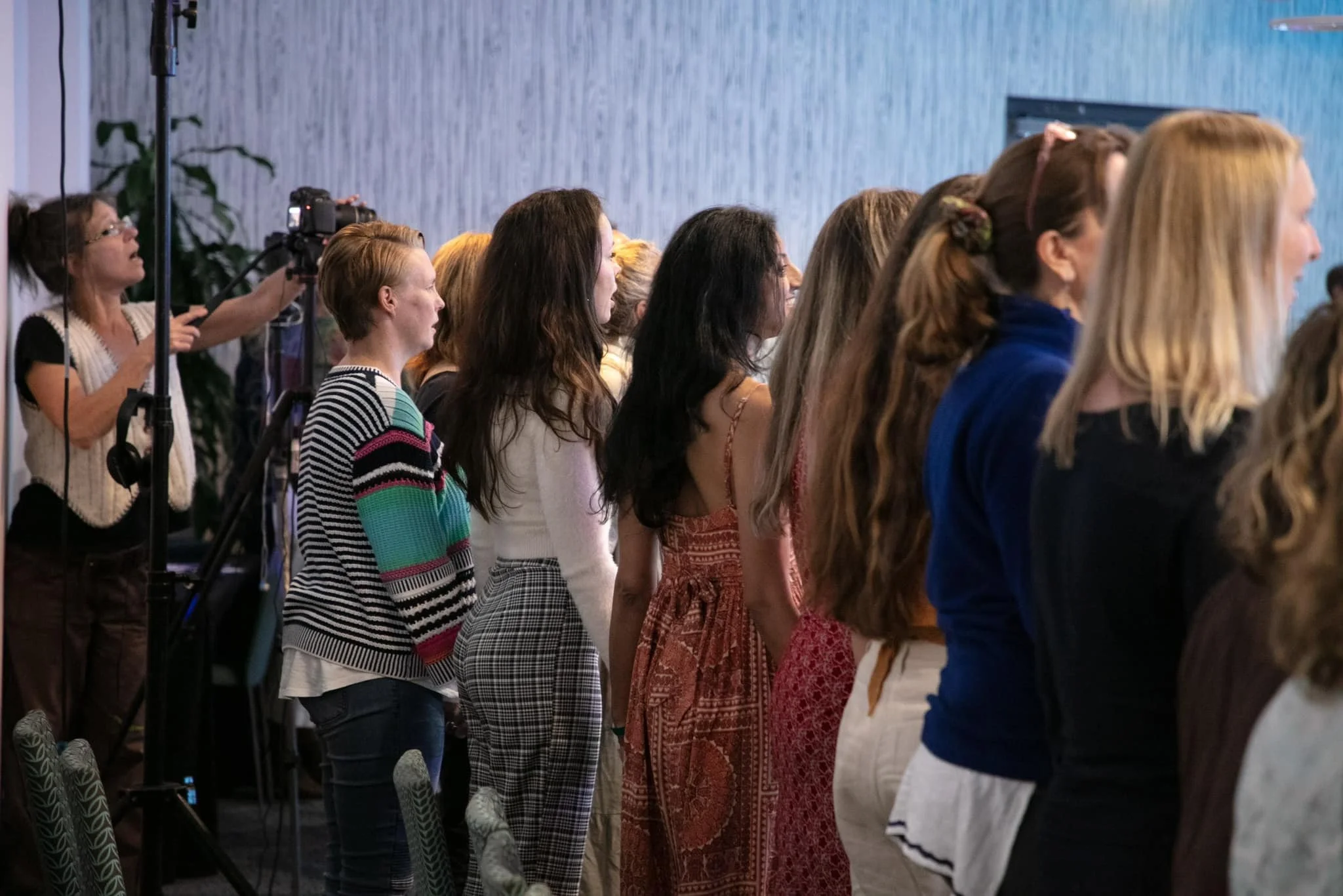 Women attending an indoor conference or seminar, standing and facing forward, with a woman recording the event on a camera in the background.