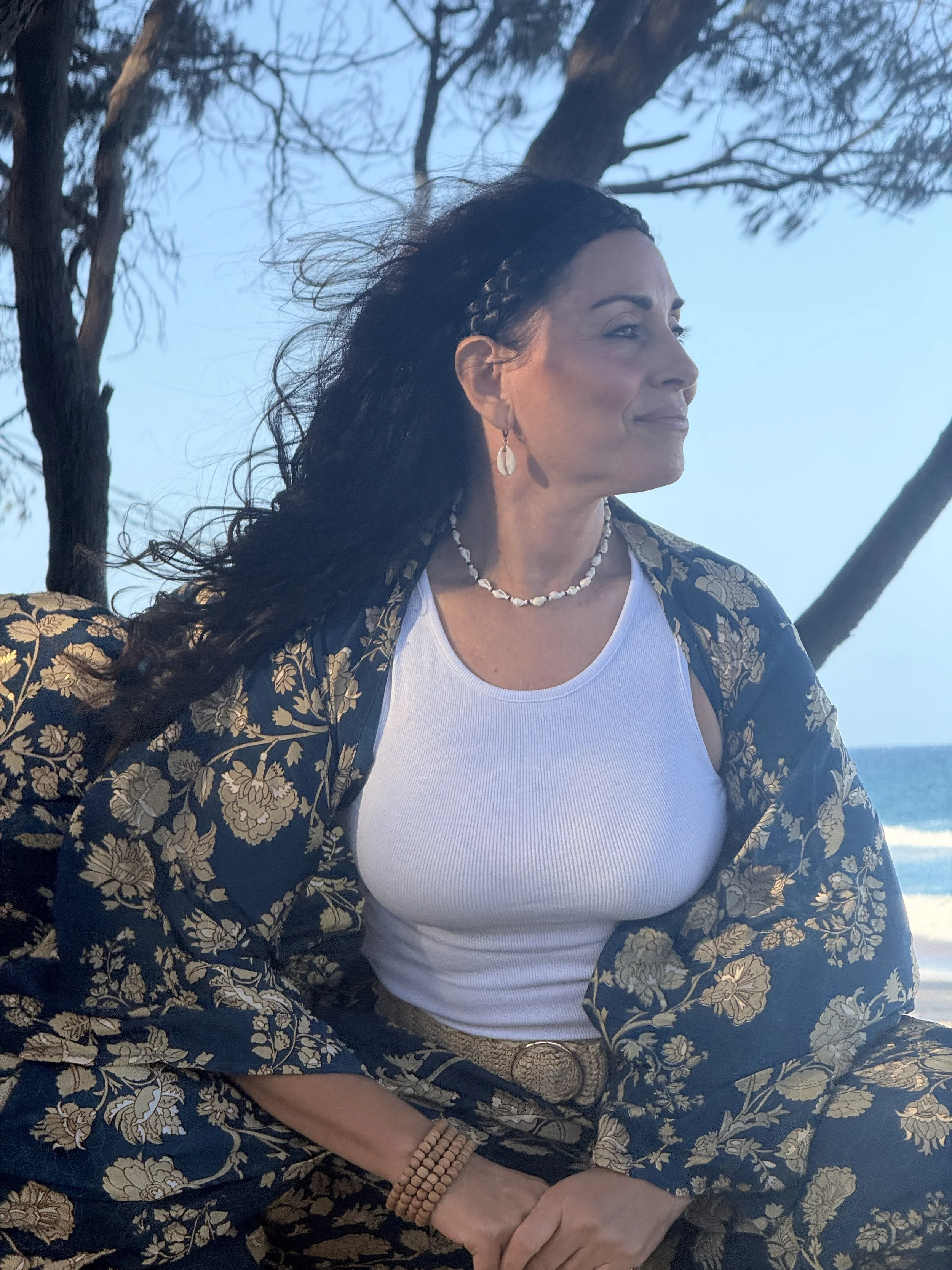 A woman with dark, wavy hair sitting outdoors near the beach, wearing a white tank top, floral patterned jacket, shell necklace, and earrings, with trees and the ocean in the background.