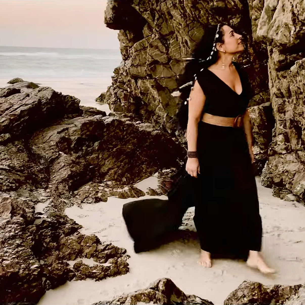 A woman walking barefoot on a sandy beach, surrounded by large rocks, with the ocean in the background.