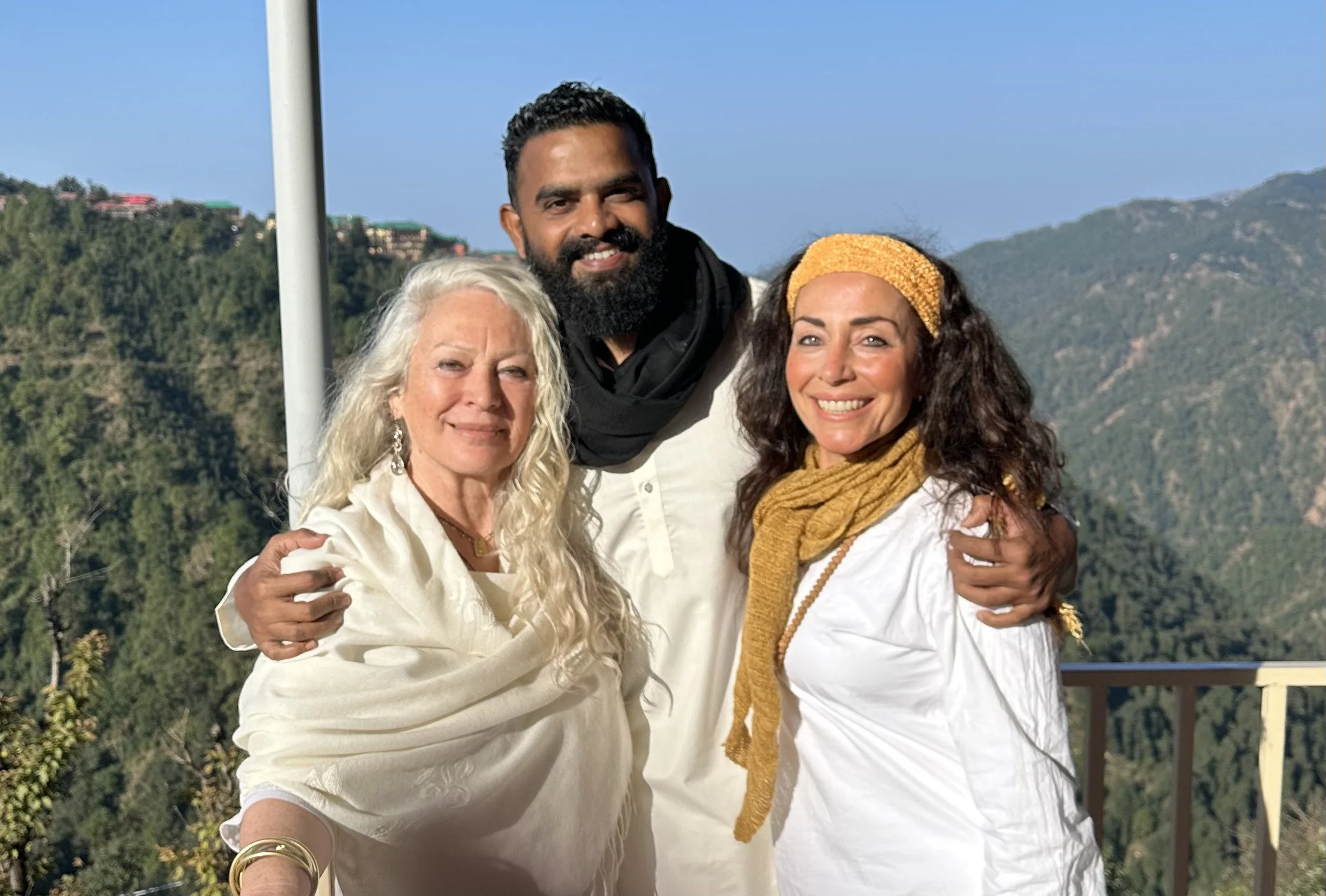 Three people, two women and one man, smiling and posing outdoors on a balcony with a mountain landscape in the background. The woman on the left has long, wavy white hair and is wearing a cream-colored shawl. The man in the middle has dark, curly hair, a full beard, and is wearing a white shirt with a black scarf. The woman on the right has dark, curly hair, a yellow headband, a musty-colored scarf, and a white shirt.