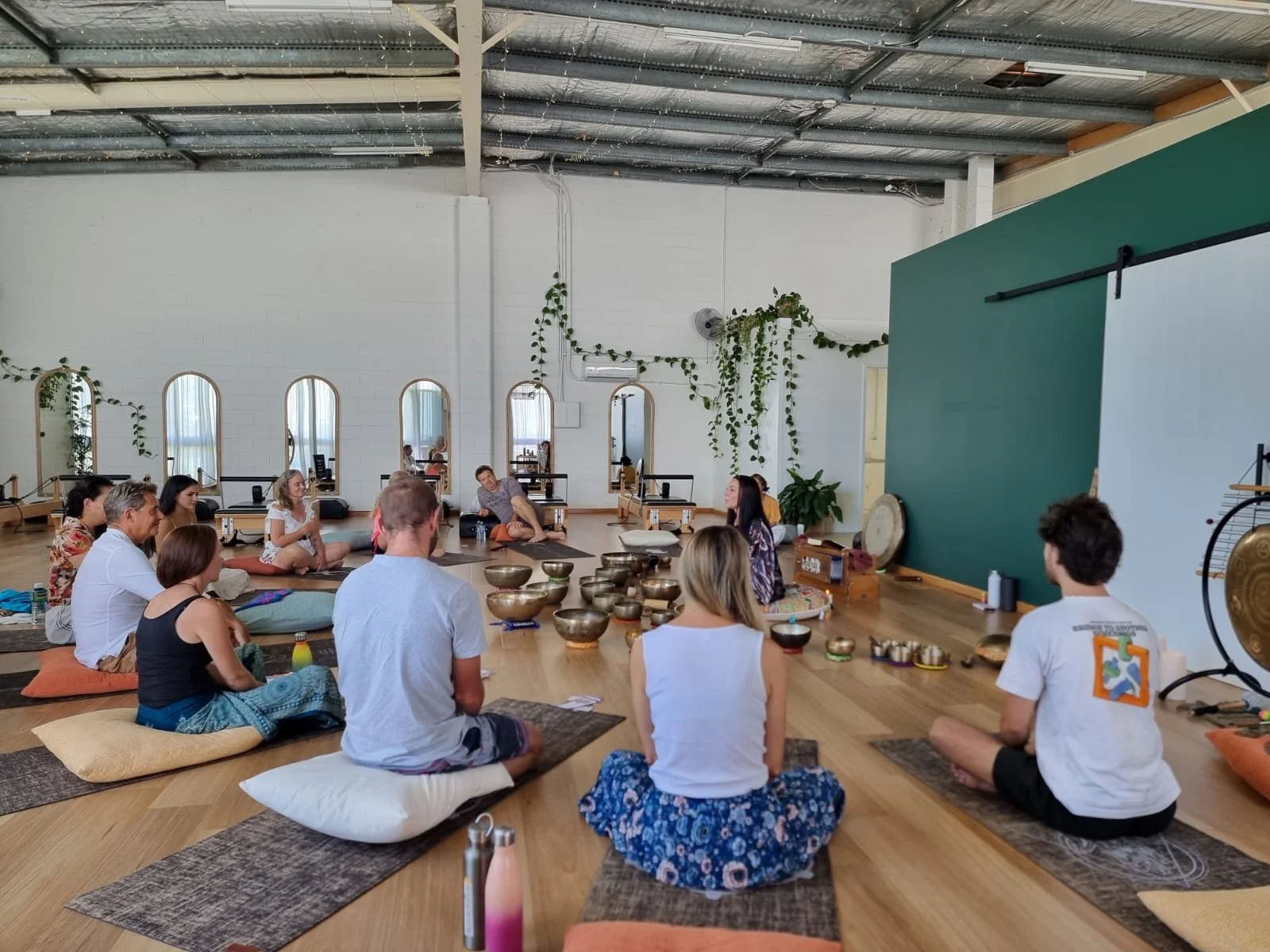 Group of people participating in a sound healing or meditation session in a spacious room with wooden floors, cushions, and various singing bowls arranged on the floor.