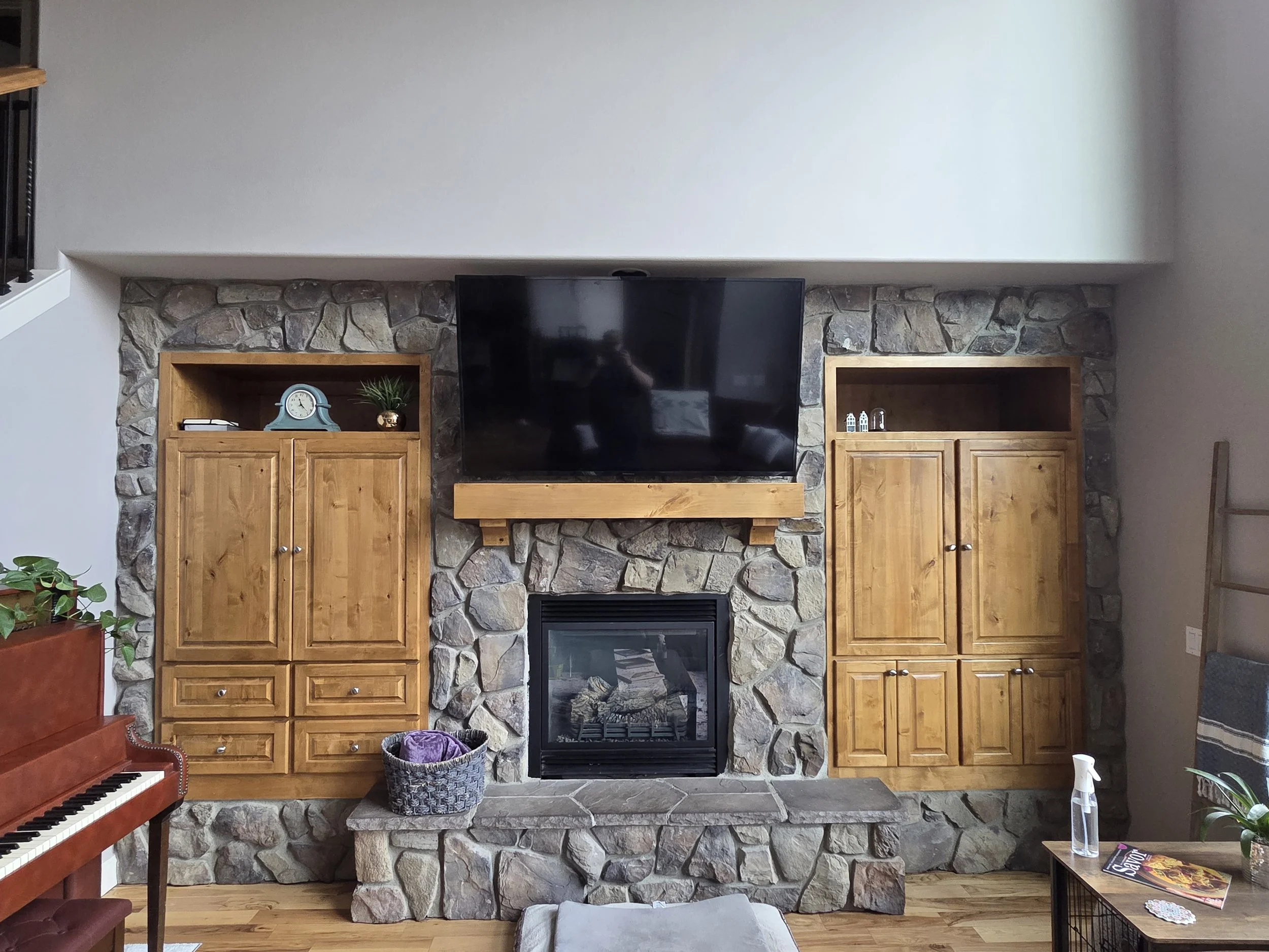 Living room with stone fireplace, wooden cabinets, flat-screen TV, purple blanket in basket, and a piano on the left.