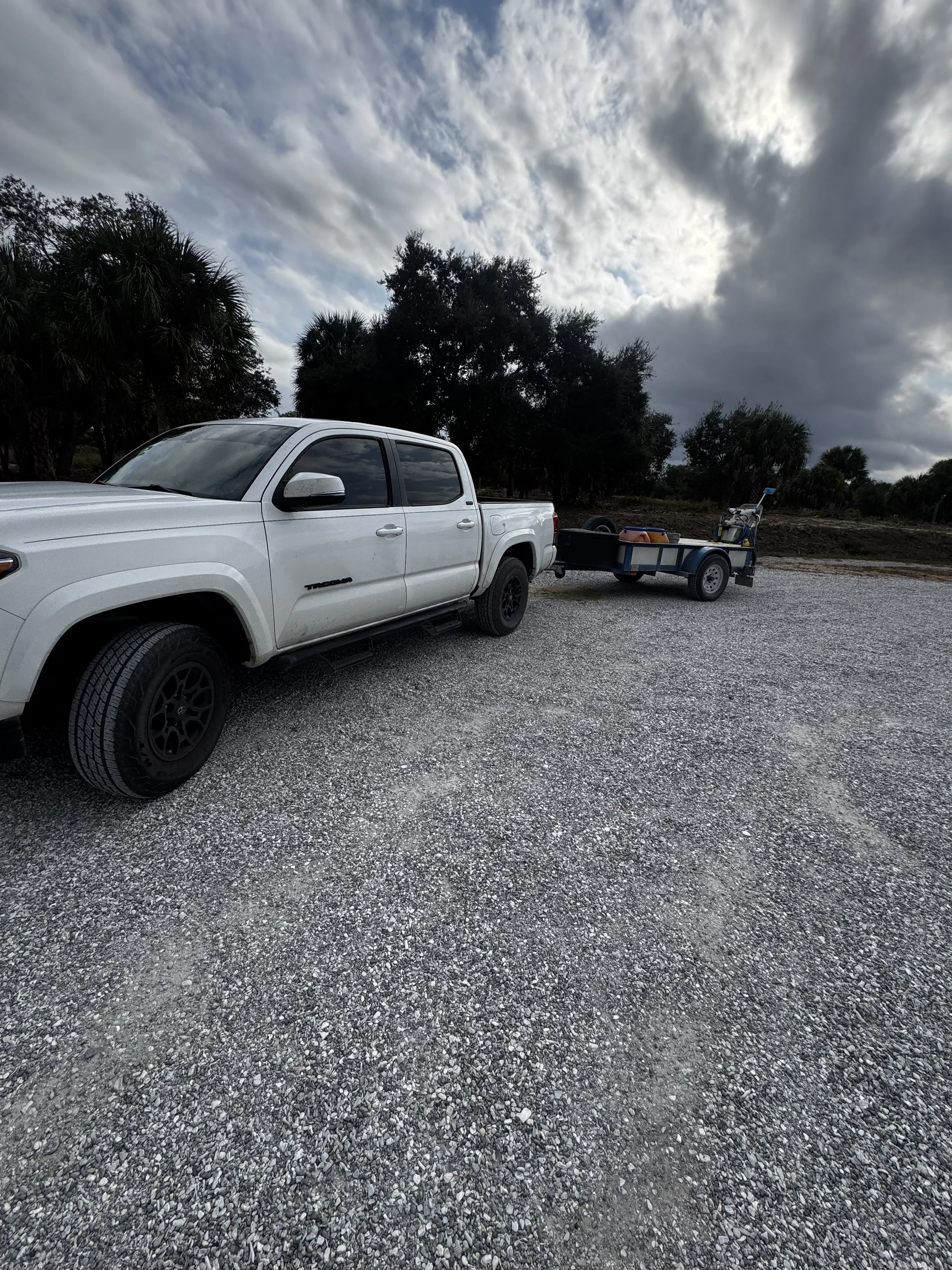 A white pickup truck parked on a gravel lot with a small trailer attached, carrying various items, and a desert landscape with trees and cloudy sky in the background.