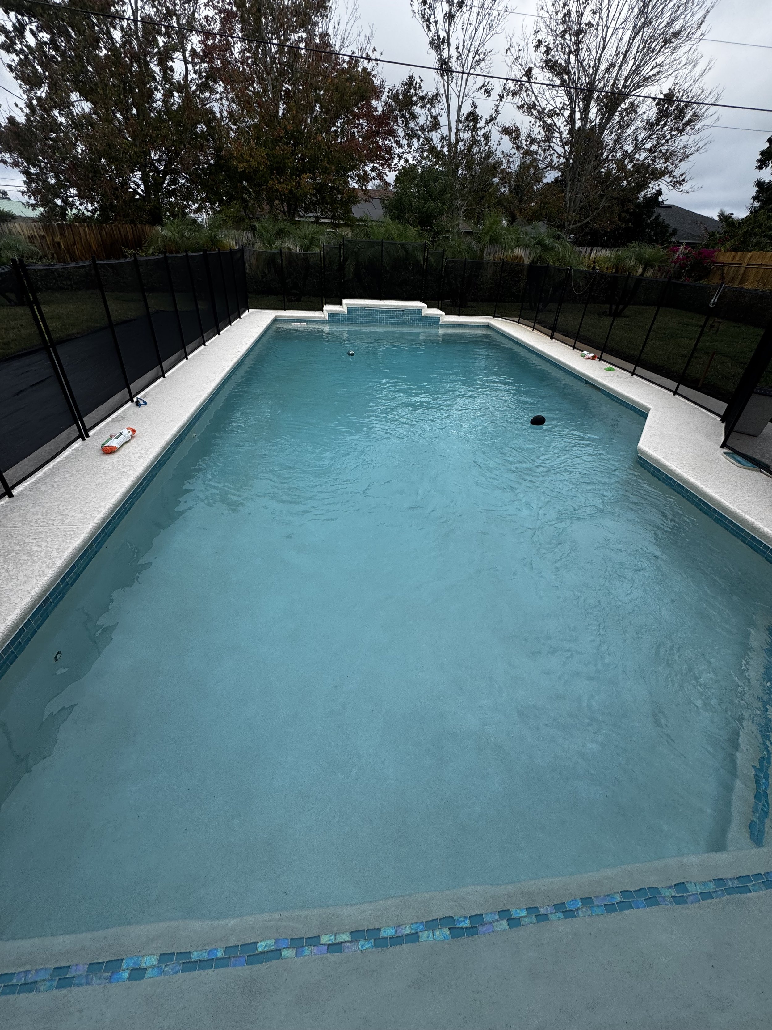 In-ground rectangular swimming pool with a light blue interior, black safety fence around it, and trees in the background on a cloudy day.