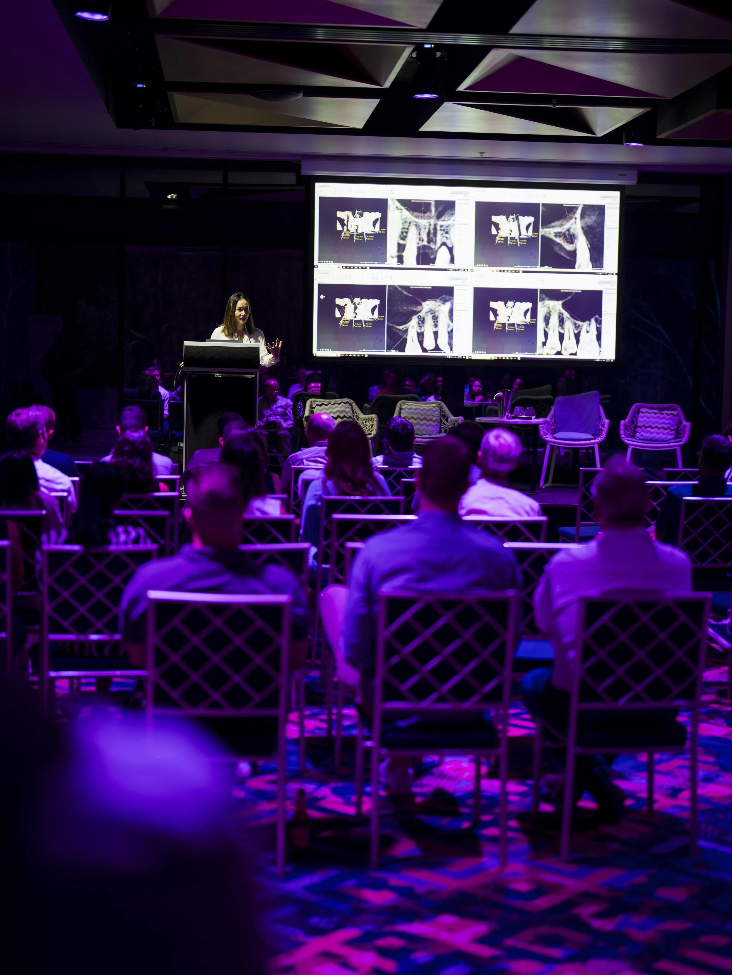 A woman is giving a presentation to an audience in a dimly lit room with purple lighting. She stands at a podium with multiple screens behind her displaying scientific images. The audience is seated in rows of chairs, attentively listening.