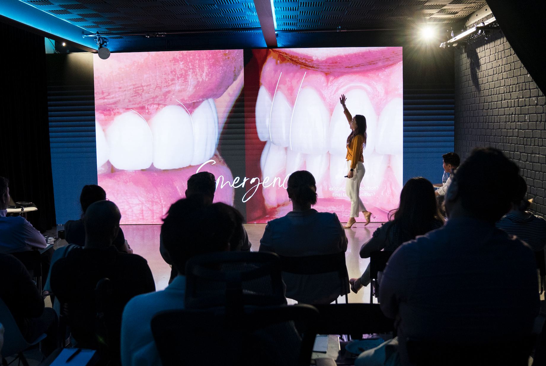 A woman giving a presentation on dental health, specifically tooth erosion, in front of a large screen showing close-up images of teeth and gums. The audience is seated in a darkened room.