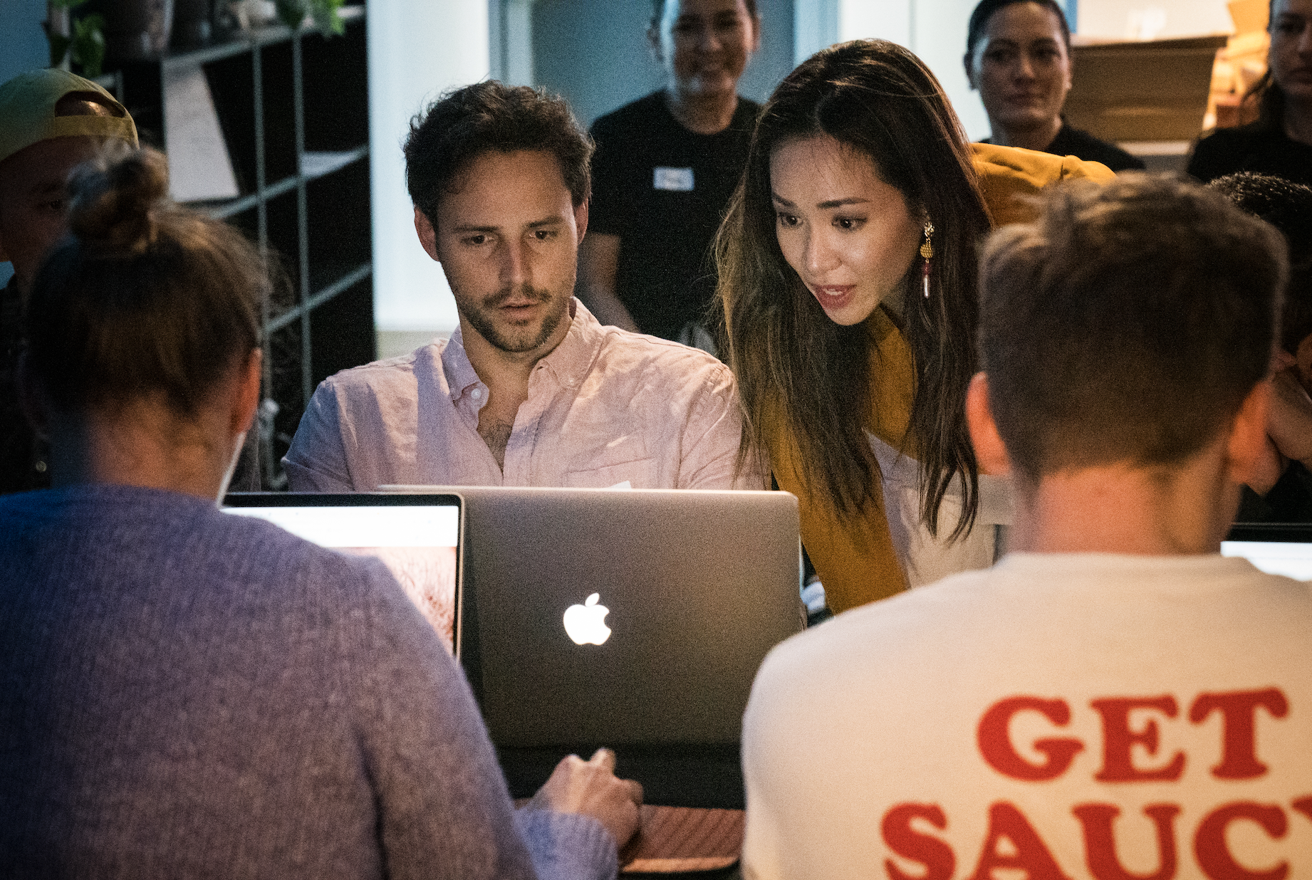 People gathered around laptops in a room, engaged in a discussion or collaboration, with some individuals looking at a screen.
