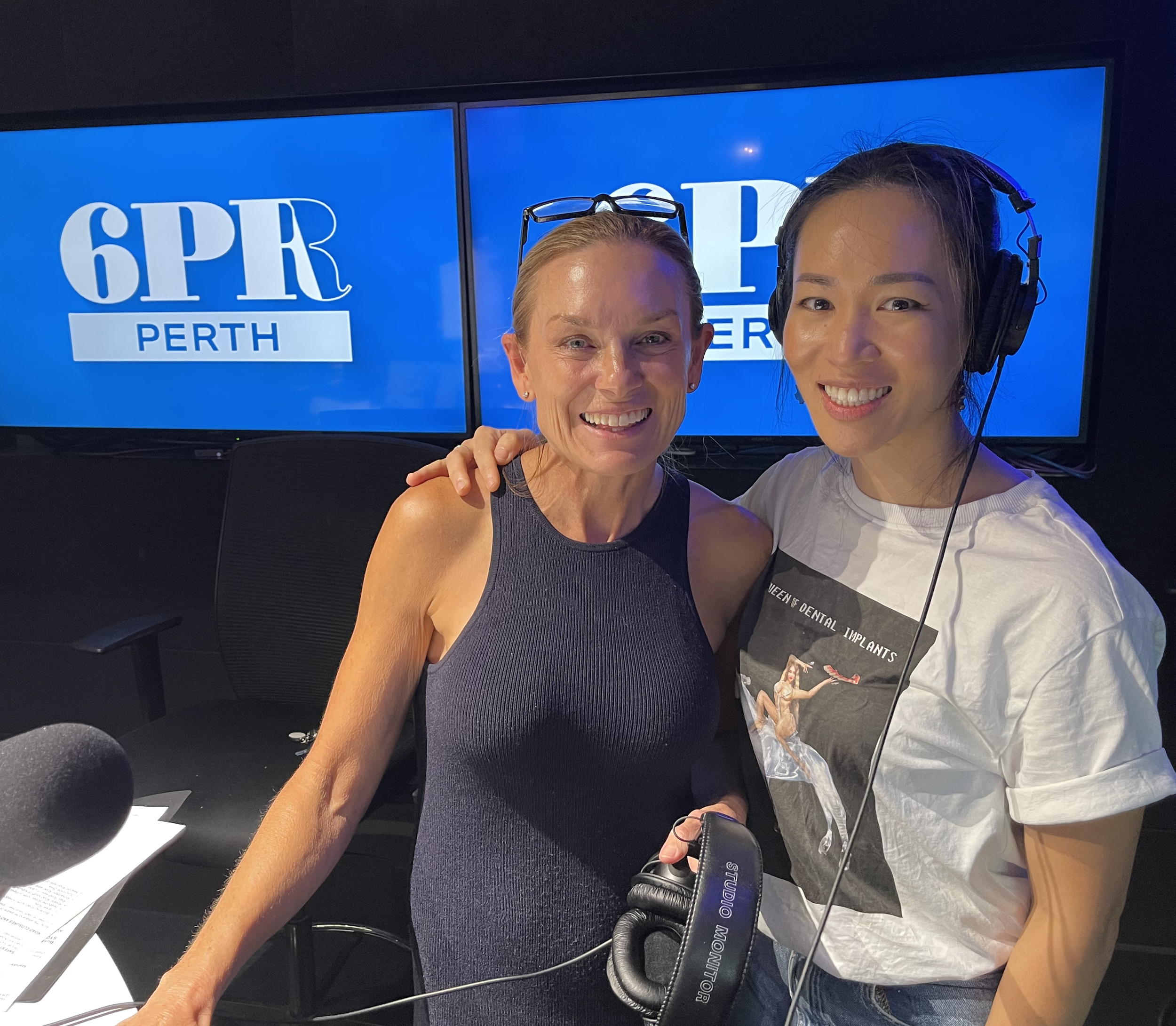 Two women smiling in a radio studio with screens that read '6PR PERTH' in the background. One is wearing a sleeveless top, and the other has a headset on and is wearing a graphic t-shirt.