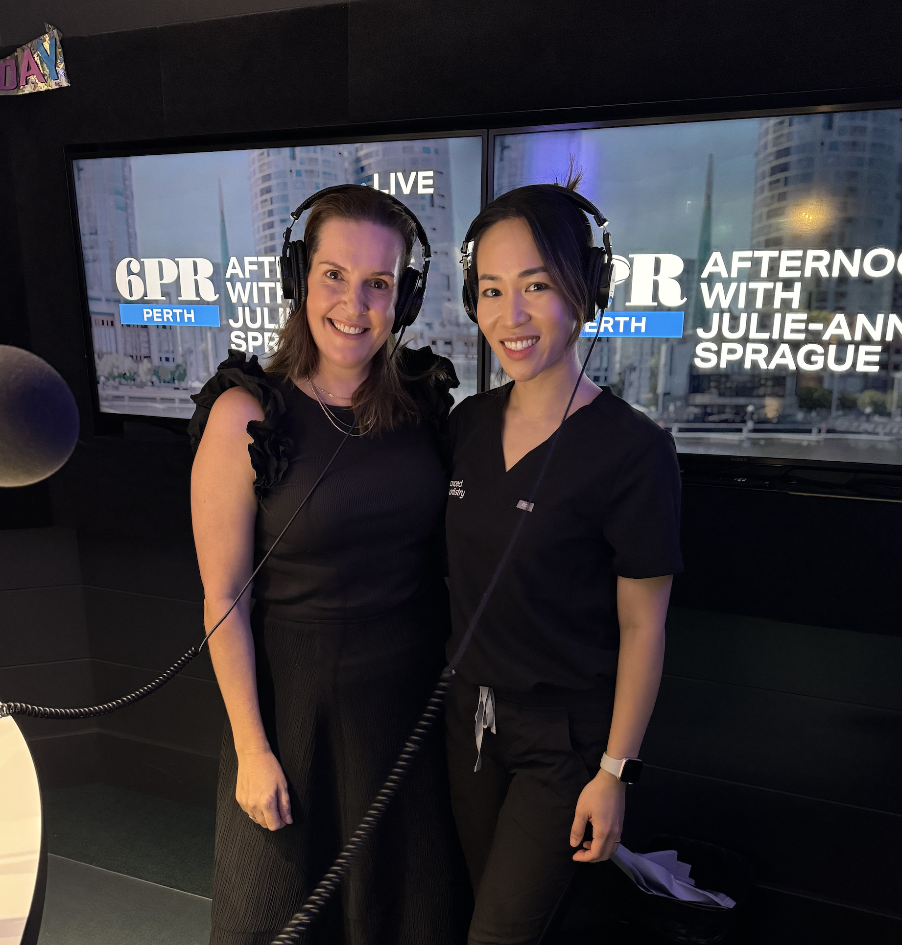 Two women standing in a recording studio, wearing large headphones, smiling at the camera, with a TV screen behind them displaying the text '6PR After With Julie-Anne Sprague Perth' and city buildings in the background.