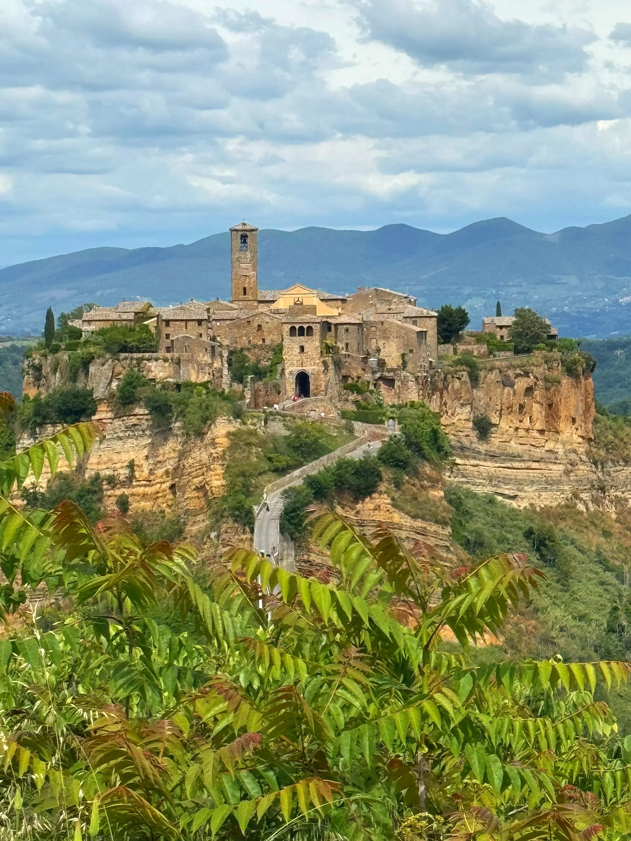 A historic hilltop village with stone buildings and a tall bell tower, surrounded by green foliage and distant mountains under a cloudy sky.