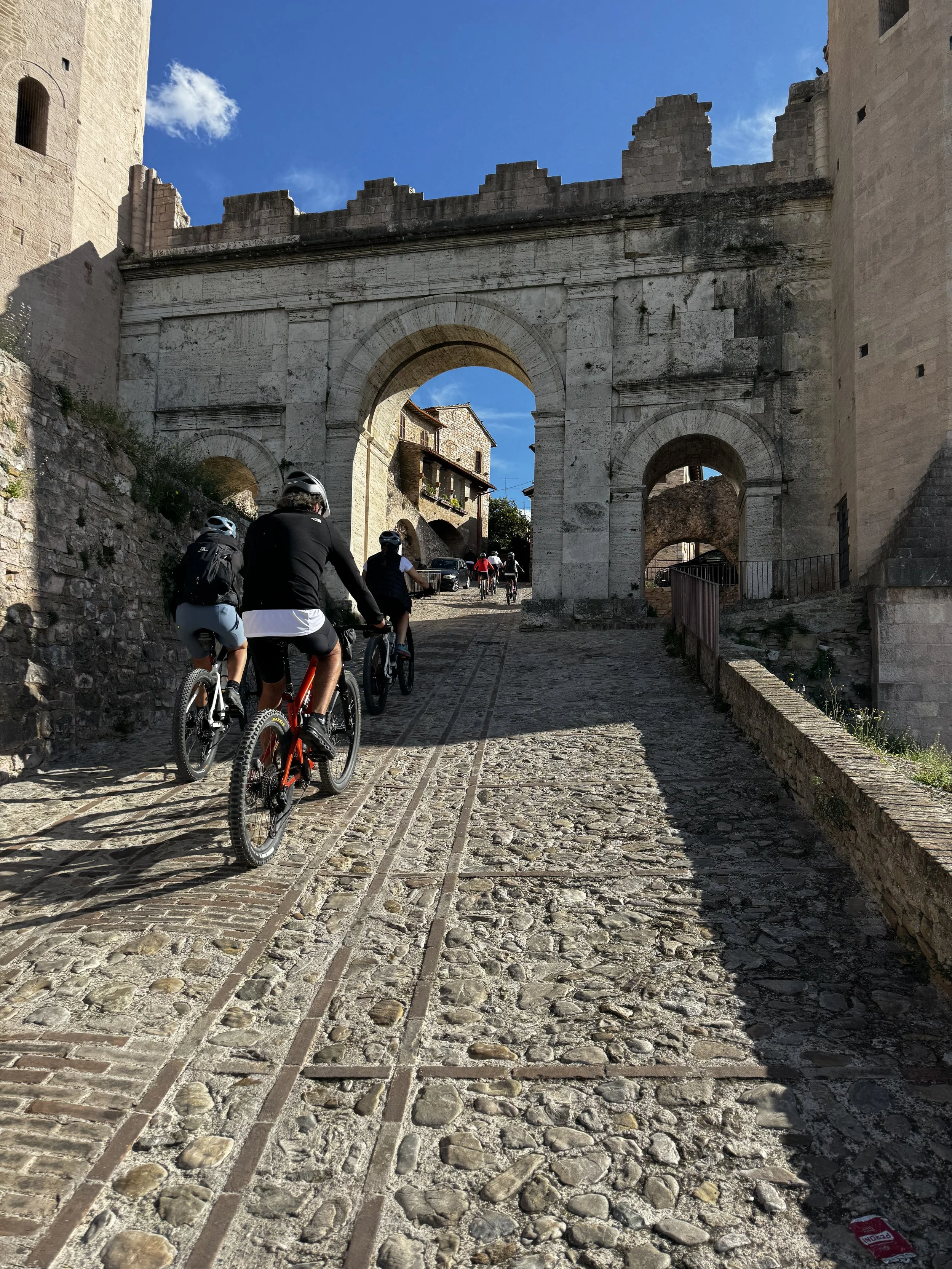 Group of people riding bicycles on a cobblestone street passing under an ancient stone archway in a historic European town.