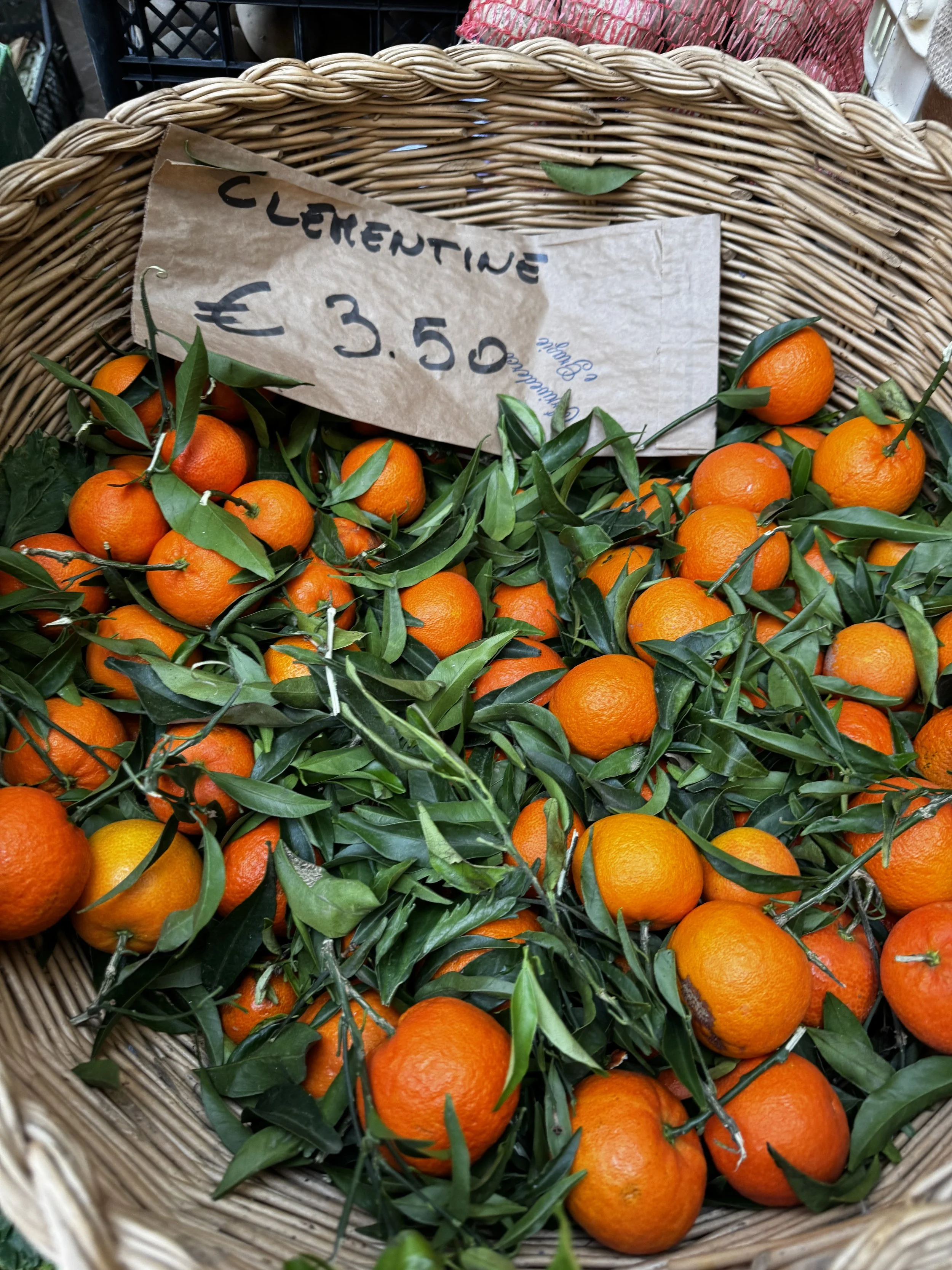 A basket of fresh clementines with green leaves attached, and a handwritten sign indicating the price of €3.50.