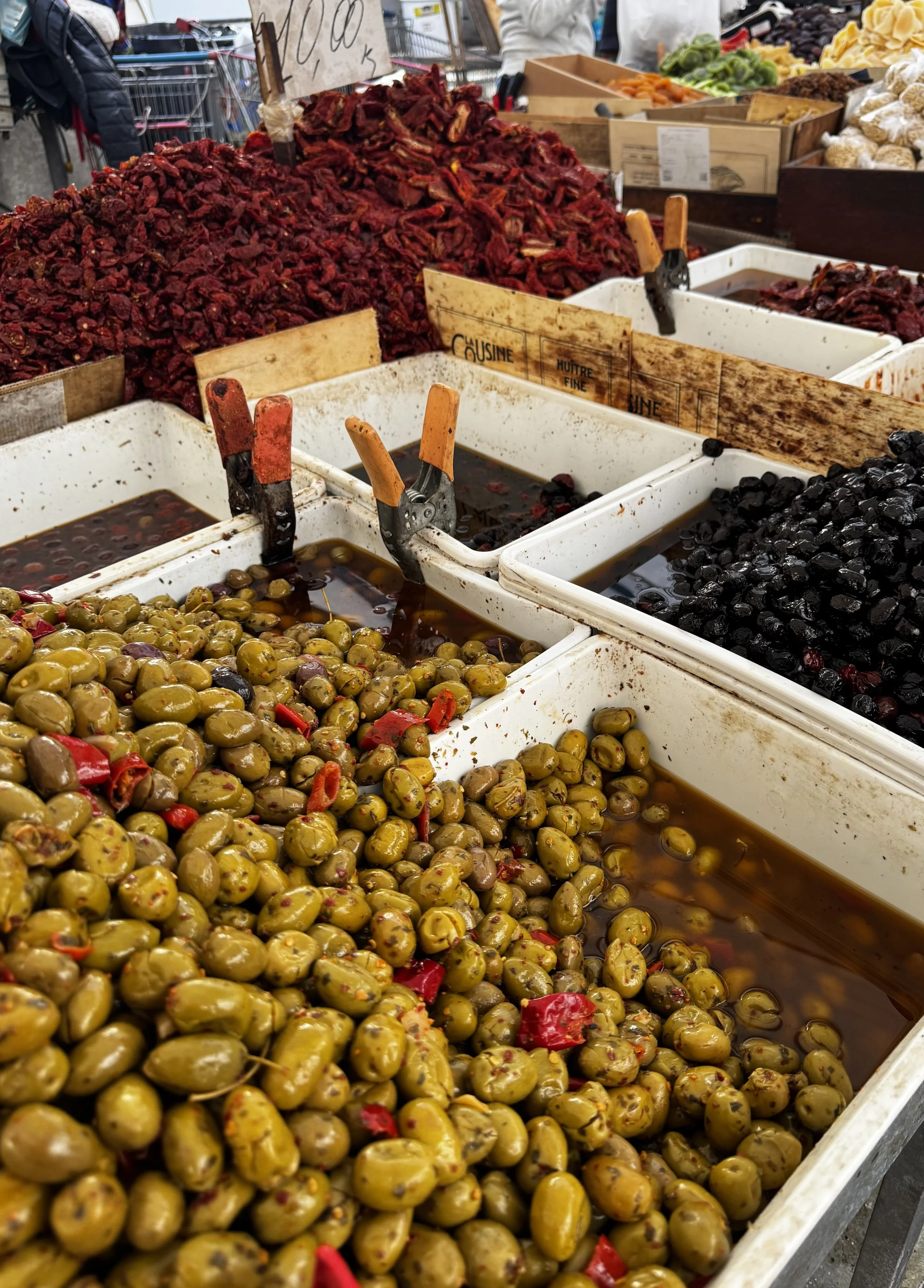 Olives at an outdoor market, with various types including green, black, and red peppers, stored in large white containers with some liquids and tongs for serving.