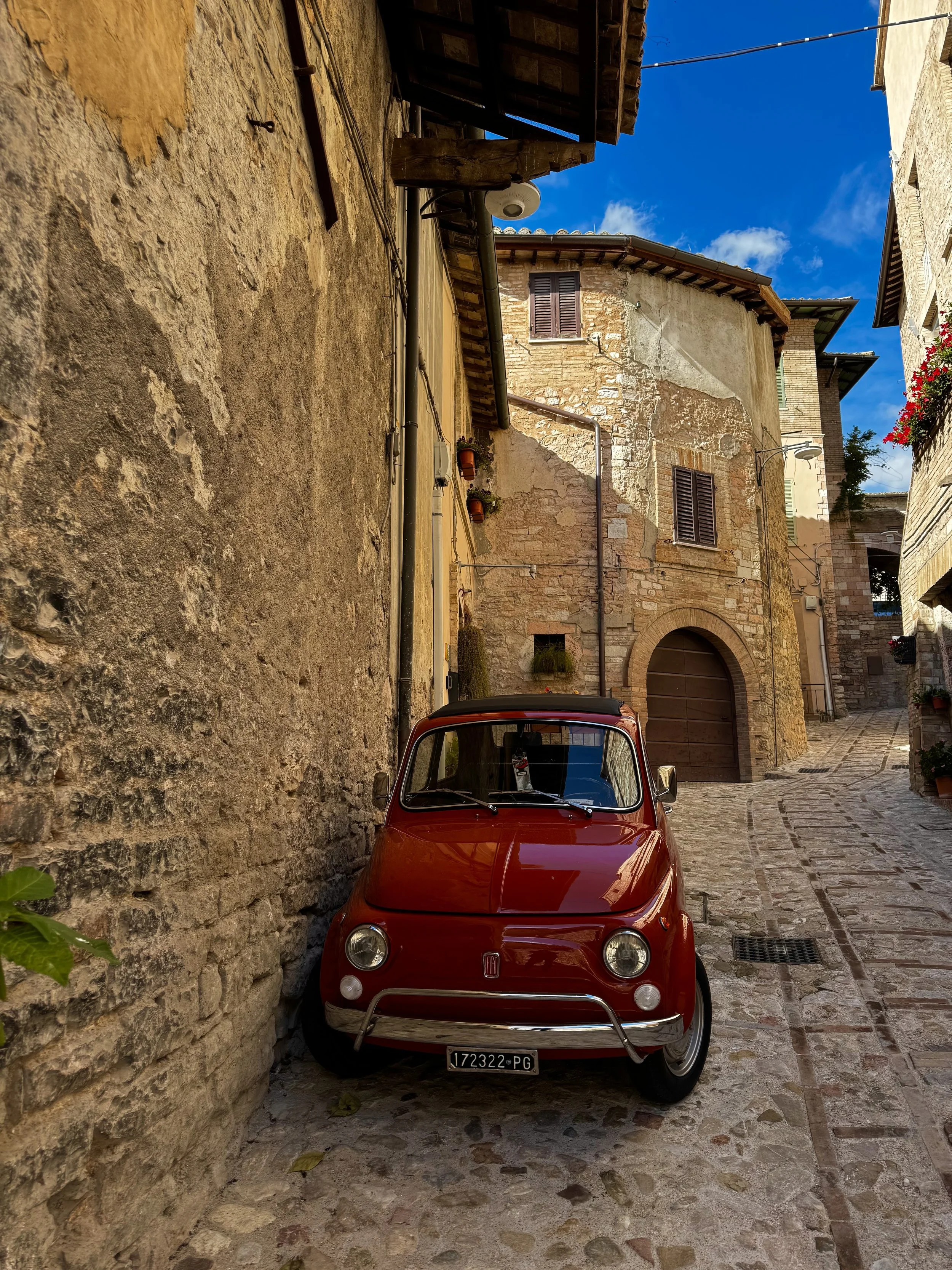 A small red vintage Fiat car parked on a cobblestone street in a historic European town with stone buildings and wooden shutters.