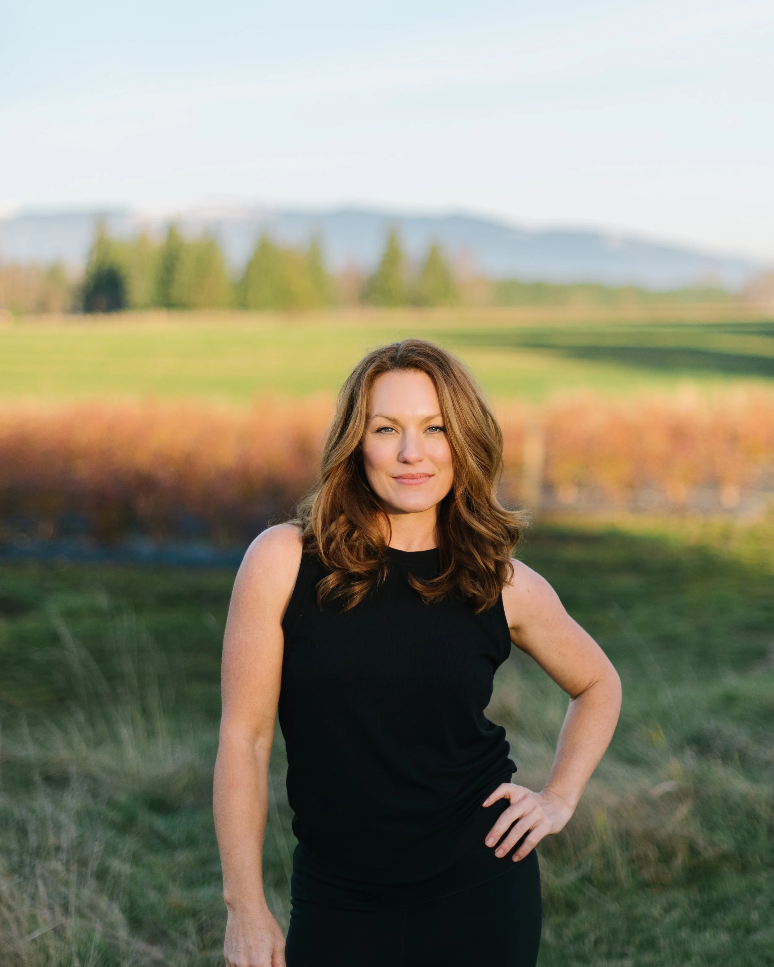 A woman with wavy, shoulder-length brown hair wearing a sleeveless black top, standing outdoors in a rural landscape with green fields, trees, and distant mountains in the background.