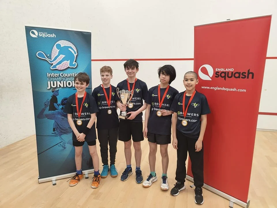 Group of five young boys standing in front of banners with squash logos, holding medals and a trophy at a squash competition.