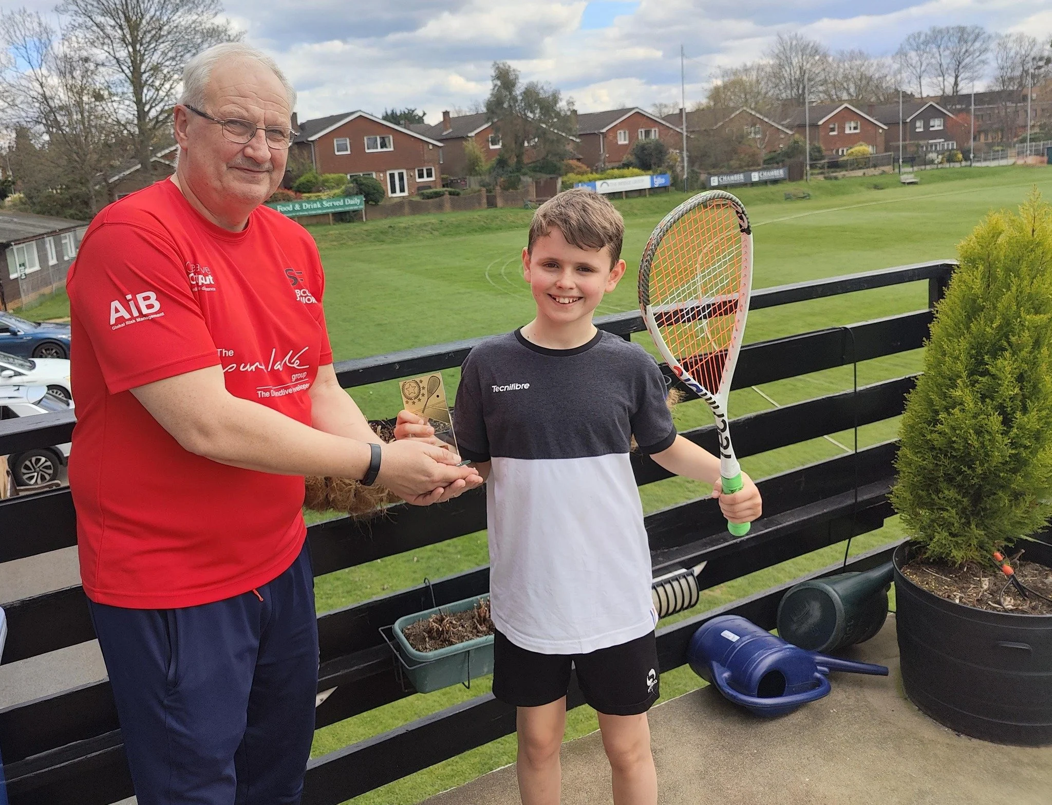 A boy holding a tennis racket smiling while receiving a trophy from an older man on a balcony overlooking a green sports field.