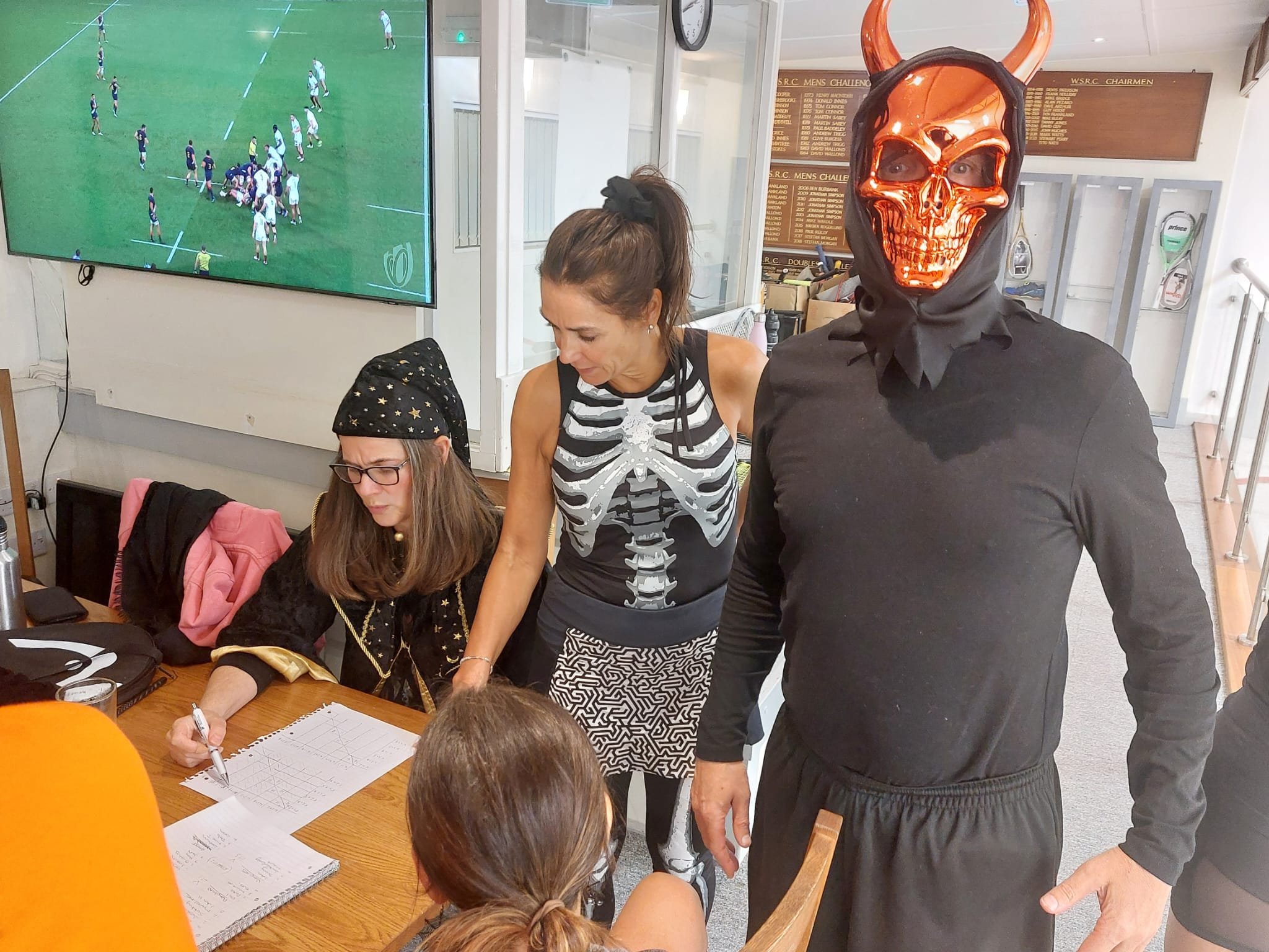 People dressed in Halloween costumes gather around a table, including a woman in a skeleton costume, a woman in a pirate costume, and a man in a devil or demon costume with a shiny skull mask and horns. A TV screen showing a rugby game is in the back