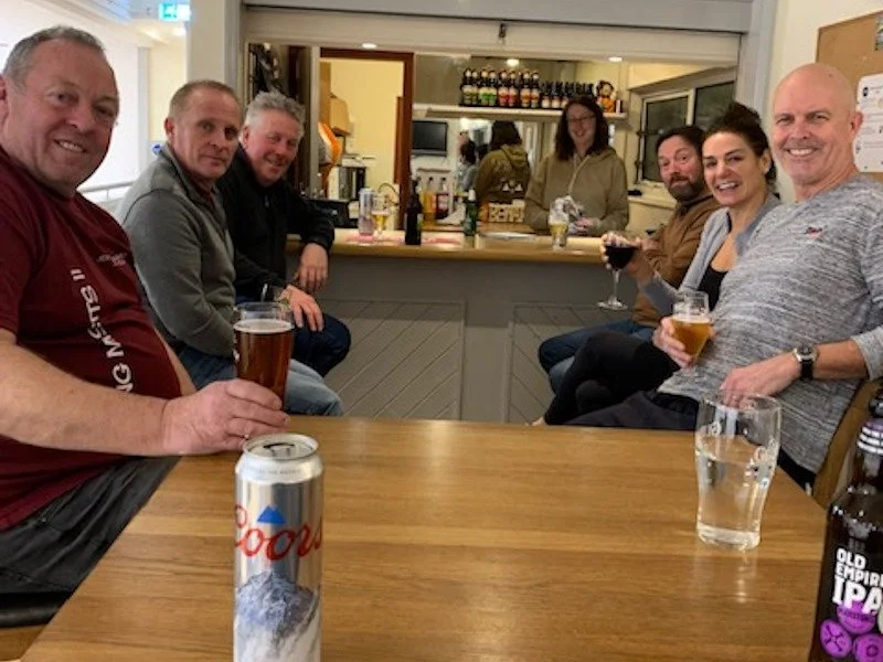 Group of seven adults sitting around a wooden table in a pub, with drinks in hand, smiling at the camera.