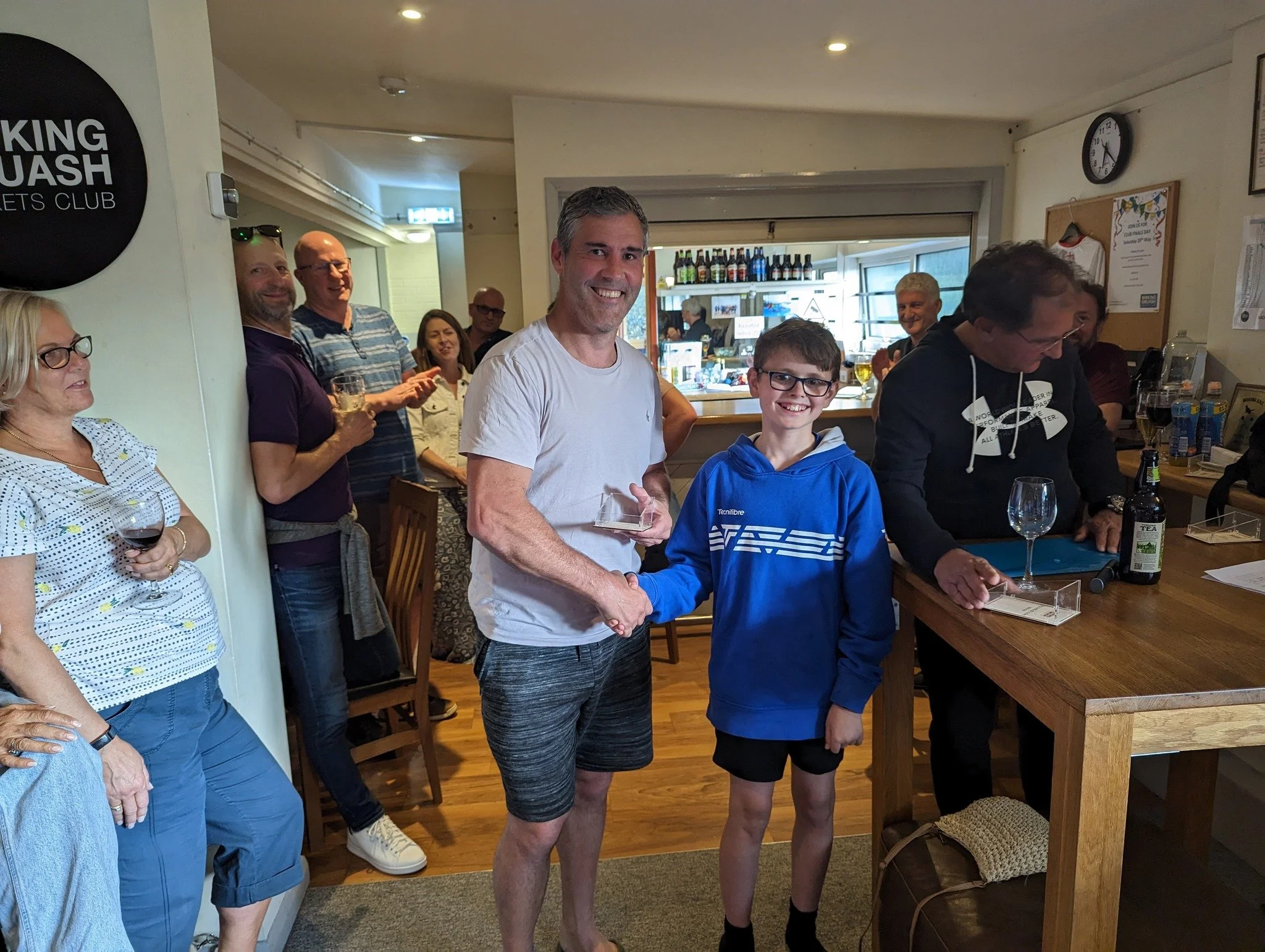 A group of people at a bar or pub, with two individuals shaking hands in the center, one holding an award. The background shows shelves with bottles, a window, and a clock. Some people are holding drinks and smiling.
