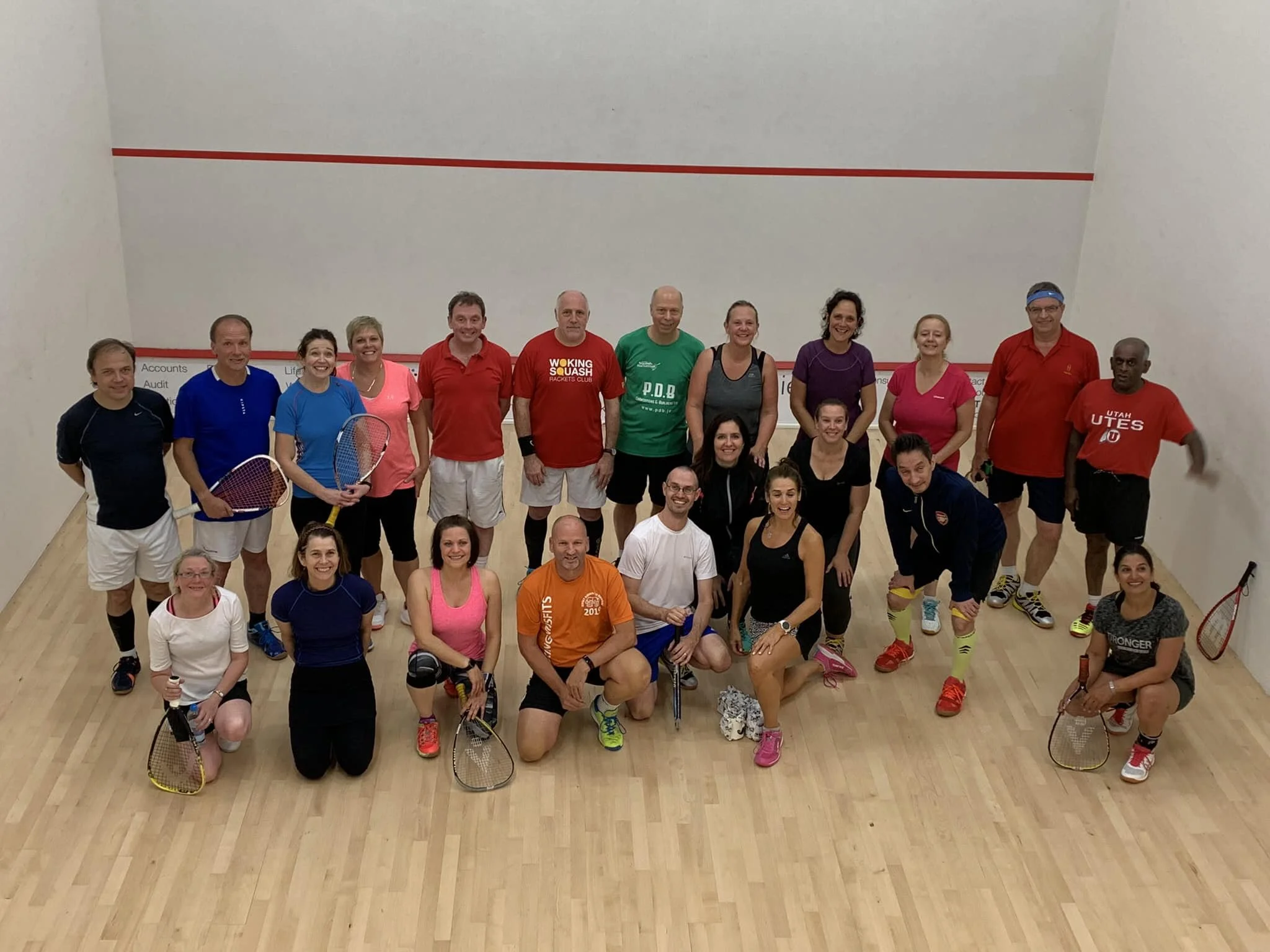 A group of 21 people on an indoor squash court, holding squash rackets, dressed in athletic clothing, smiling for a group photo.