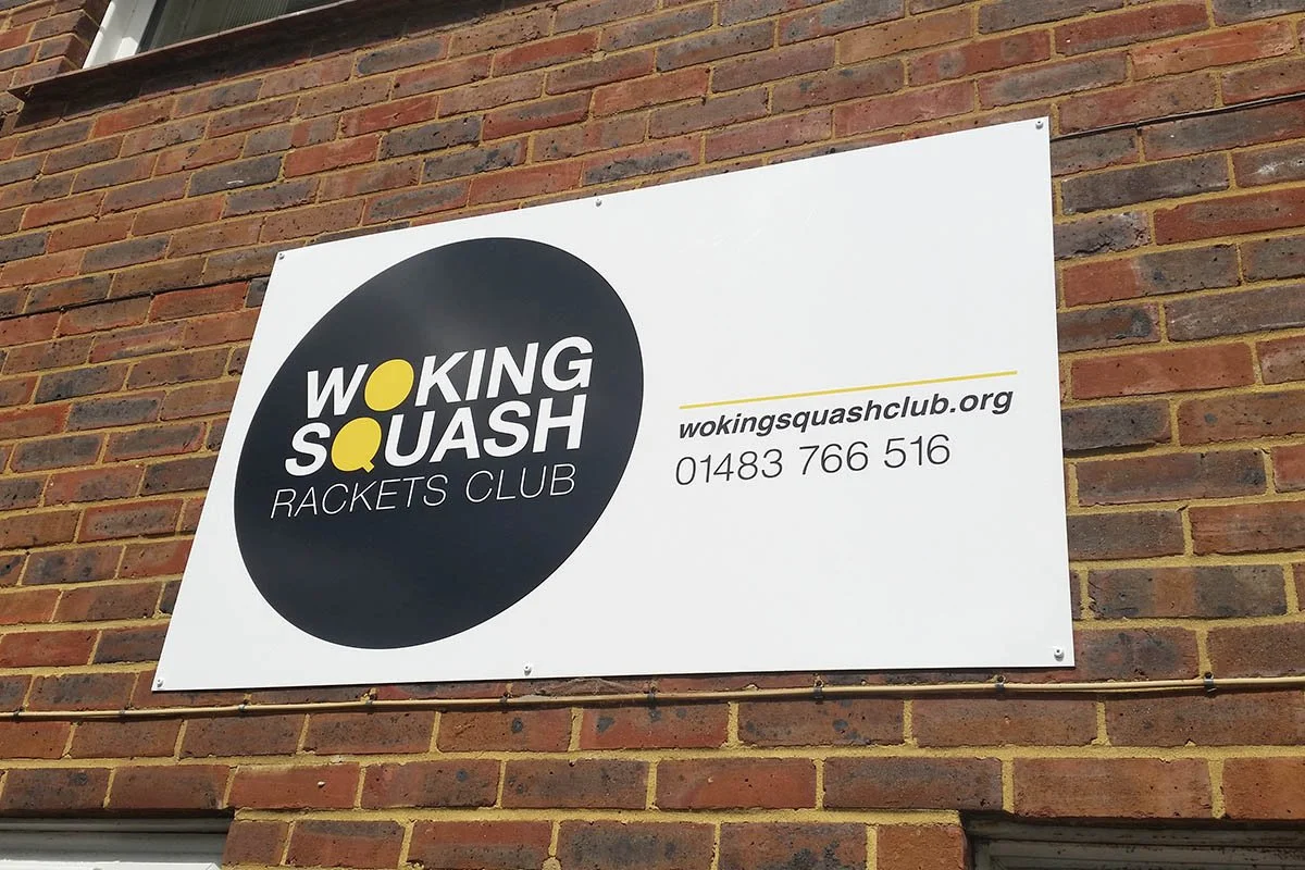 Sign for Woking Squash Rackets Club mounted on a brick wall, displaying the club's name, website, and phone number.