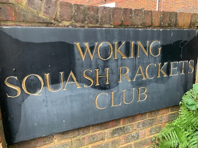 A sign on a brick wall that reads "Woking Squash Rackets Club" in gold letters.