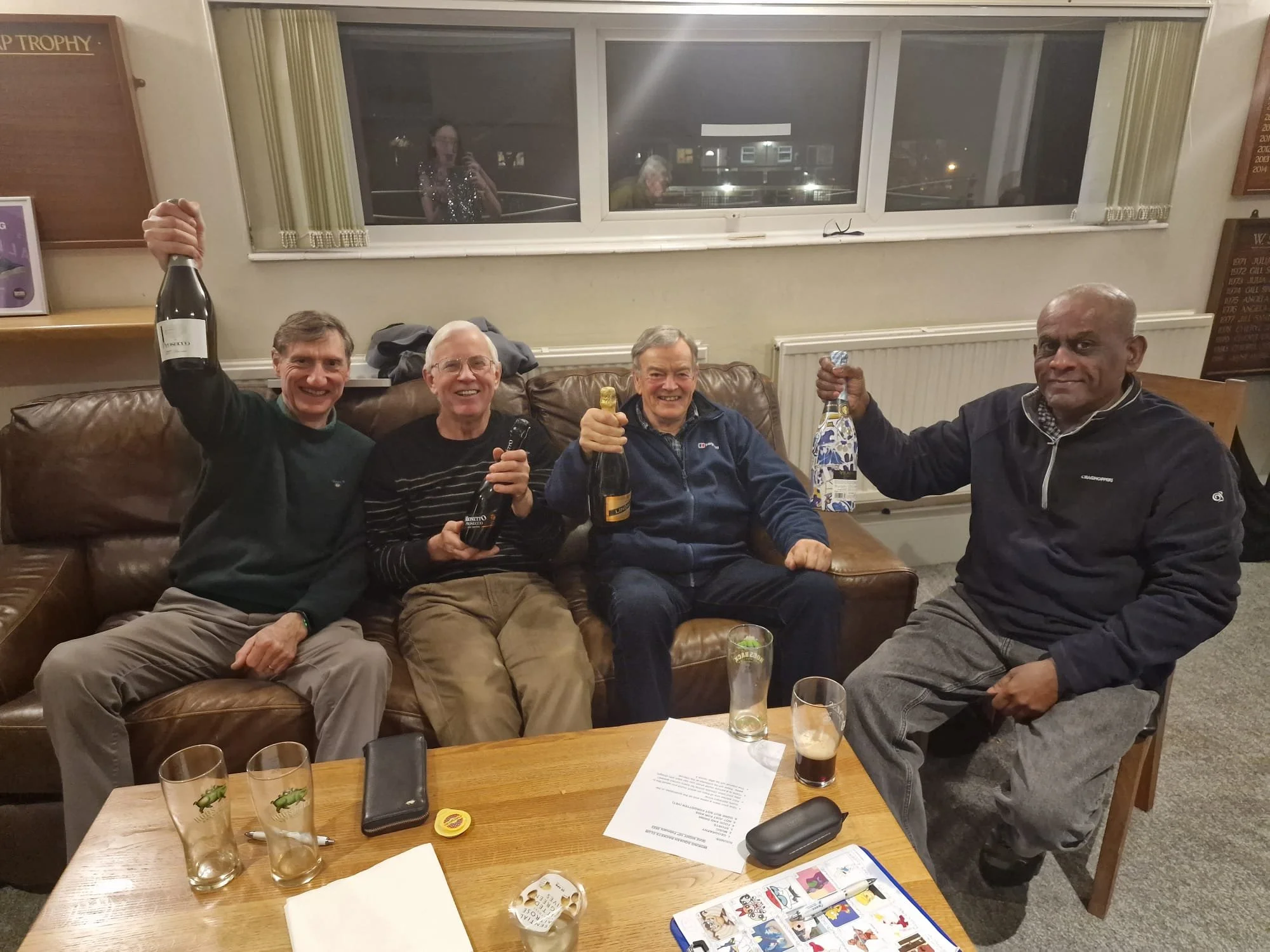 Four men sitting on a brown leather couch at a social gathering, raising bottles of beer and smiling. A coffee table in front holds empty glasses, a menu, and personal items. The room has a window with a reflection of people and lights outside.