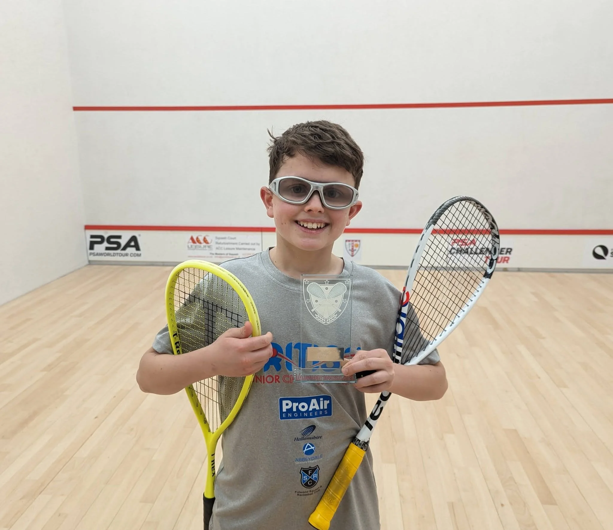 A young boy holding two squash rackets and a glass trophy, smiling in an indoor squash court.