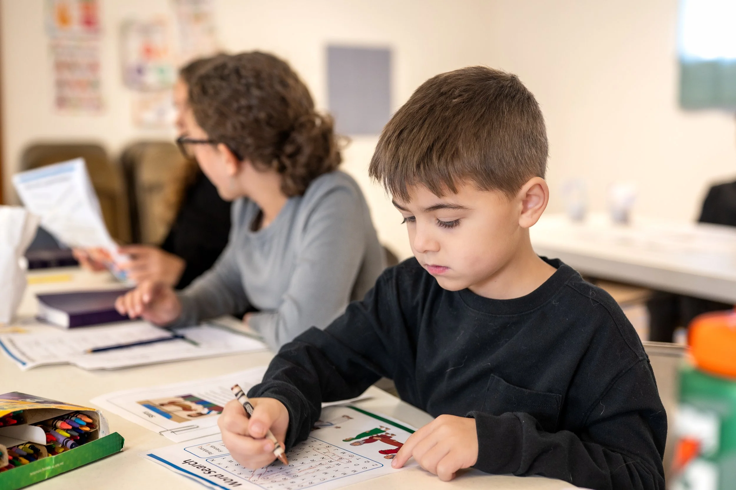A young boy with brown hair focused on writing at a classroom desk, with a woman and another student in the background.