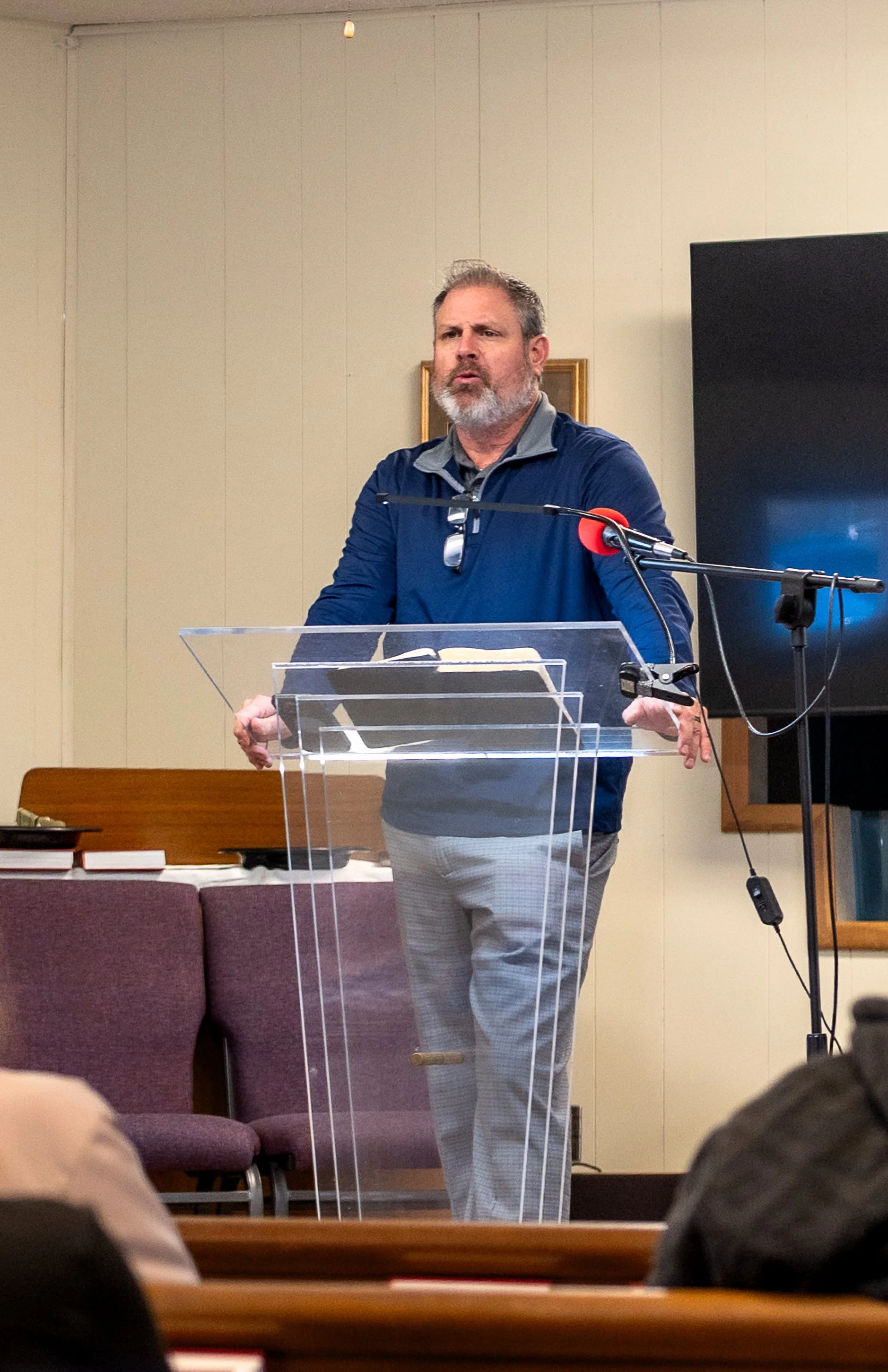 A pastor or preacher speaking at a podium with a microphone, in a room with beige walls, purple chairs, and a large screen.