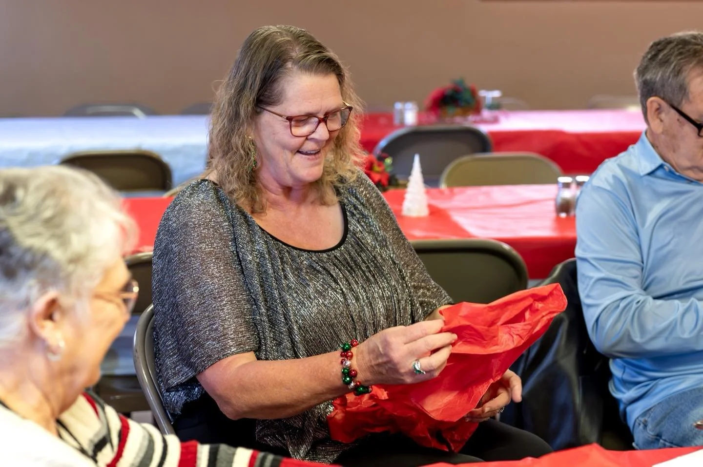 A woman with glasses and curly hair smiling while opening a red gift bag at a holiday event, seated at a table with other people.
