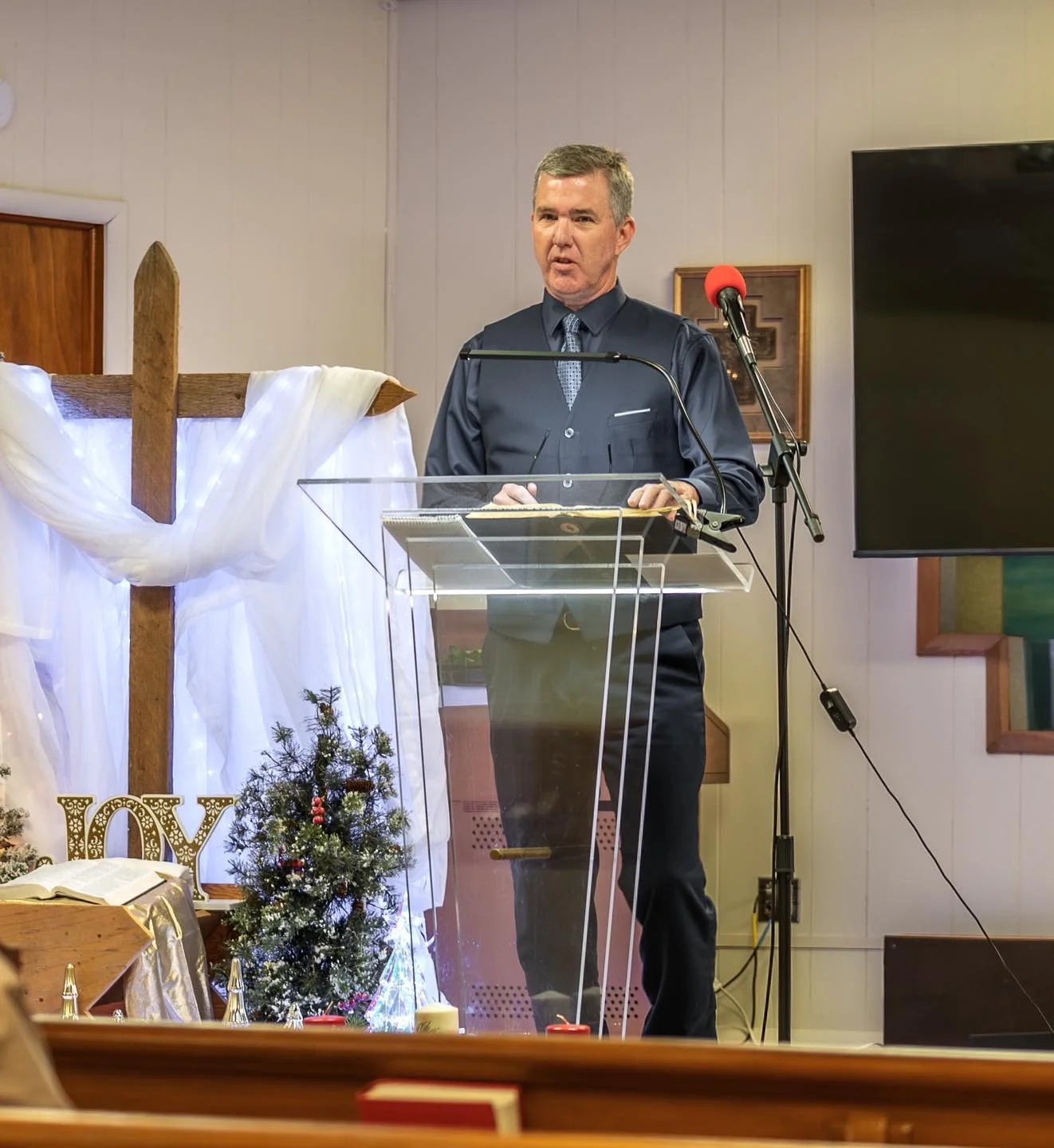 A man in a dark shirt and tie stands behind a transparent pulpit, speaking into a microphone in a decorated room with Christmas decorations, including a small Christmas tree, white drapery with a wooden cross, and gold ornaments.