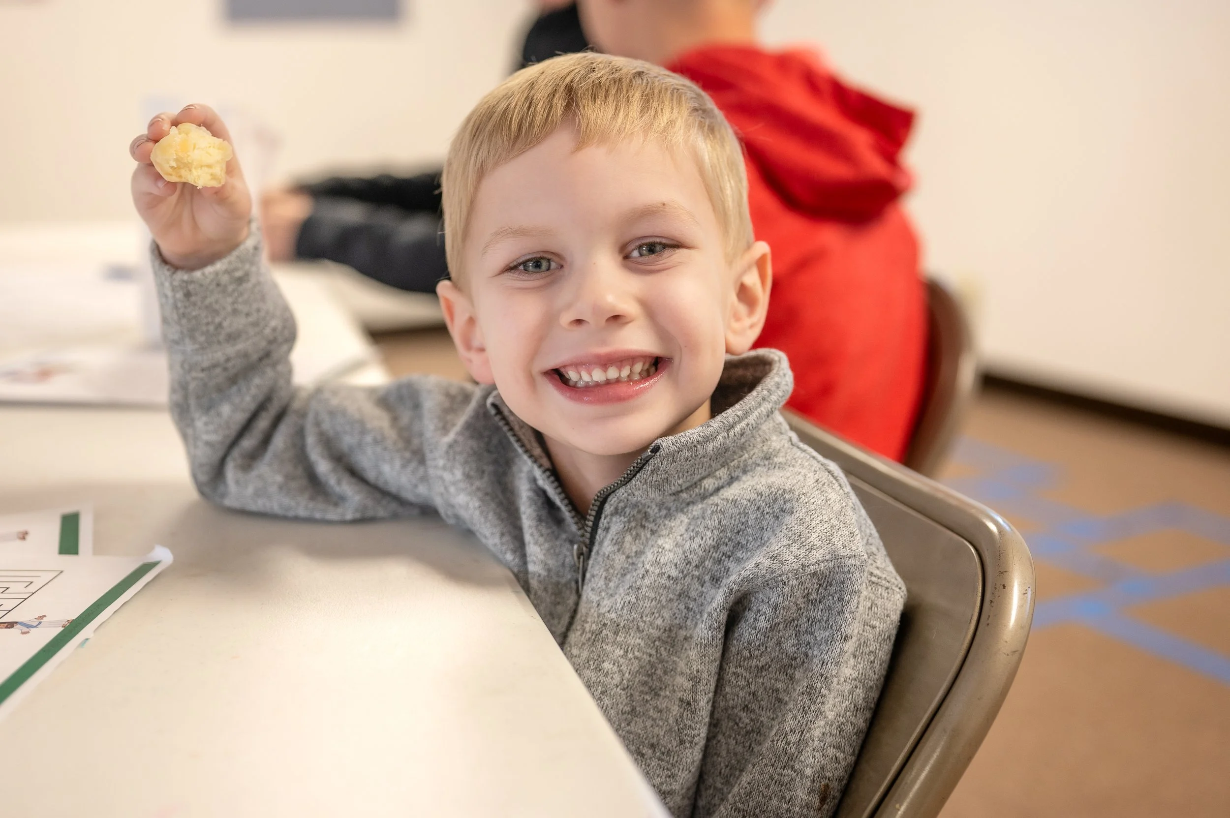 A young boy with blond hair and a gray jacket smiling and holding a piece of bread or a similar food item in his right hand. He is sitting at a table with a worksheet and other children in the background.