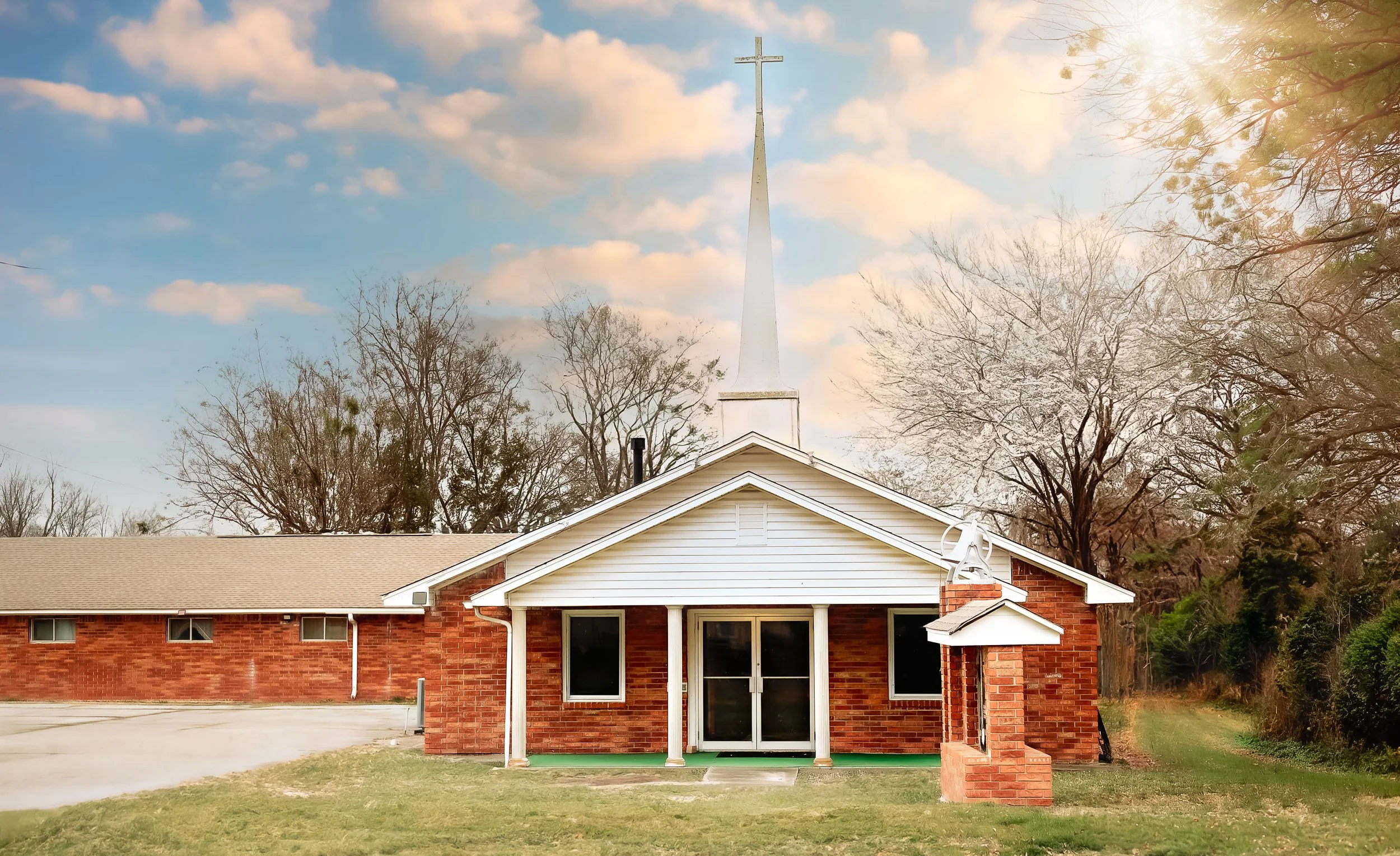 Front view of a small brick church with white trim and a steeple topped with a cross, surrounded by trees on a partly cloudy day.