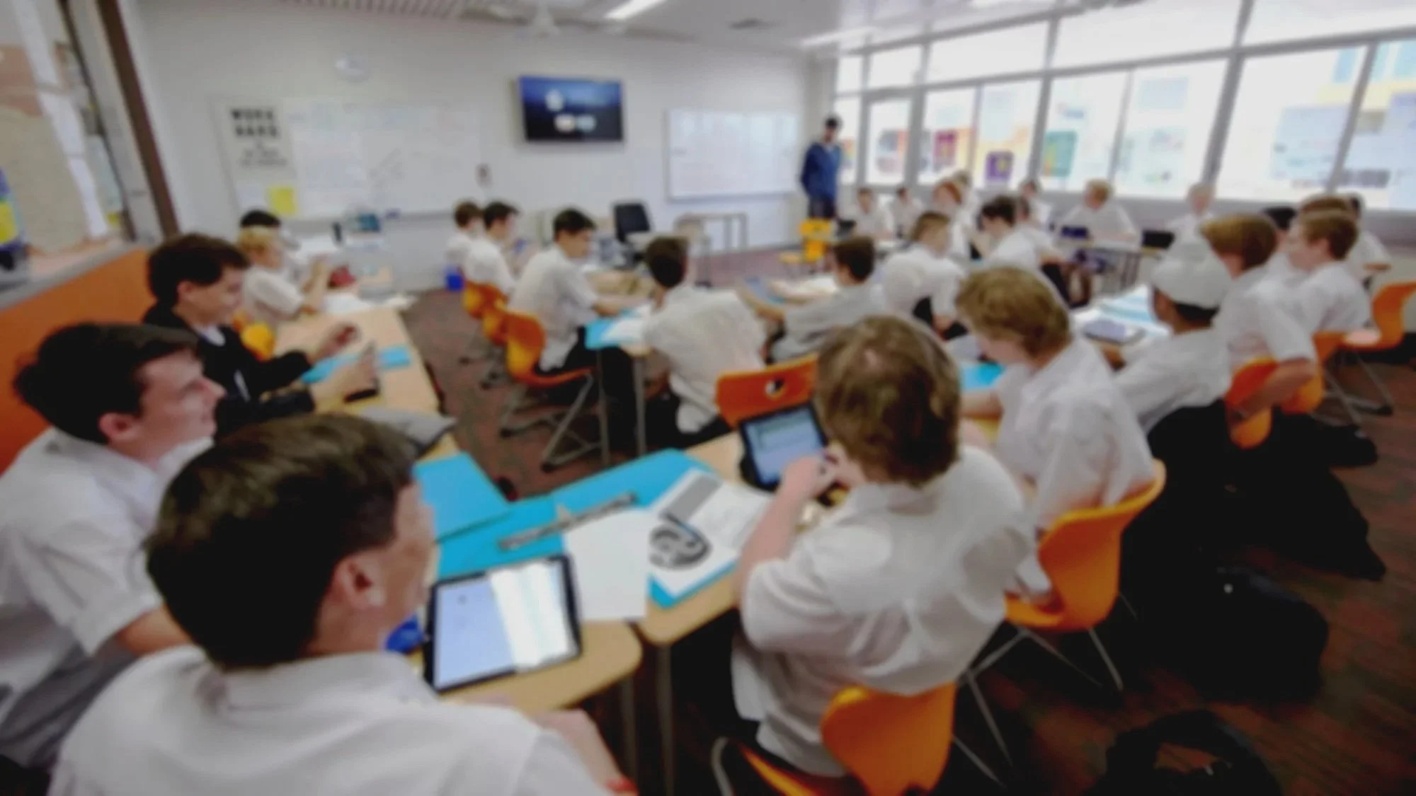 Classroom filled with students sitting at desks, working on tablets, with a teacher at the front near a whiteboard and a large window letting in daylight.