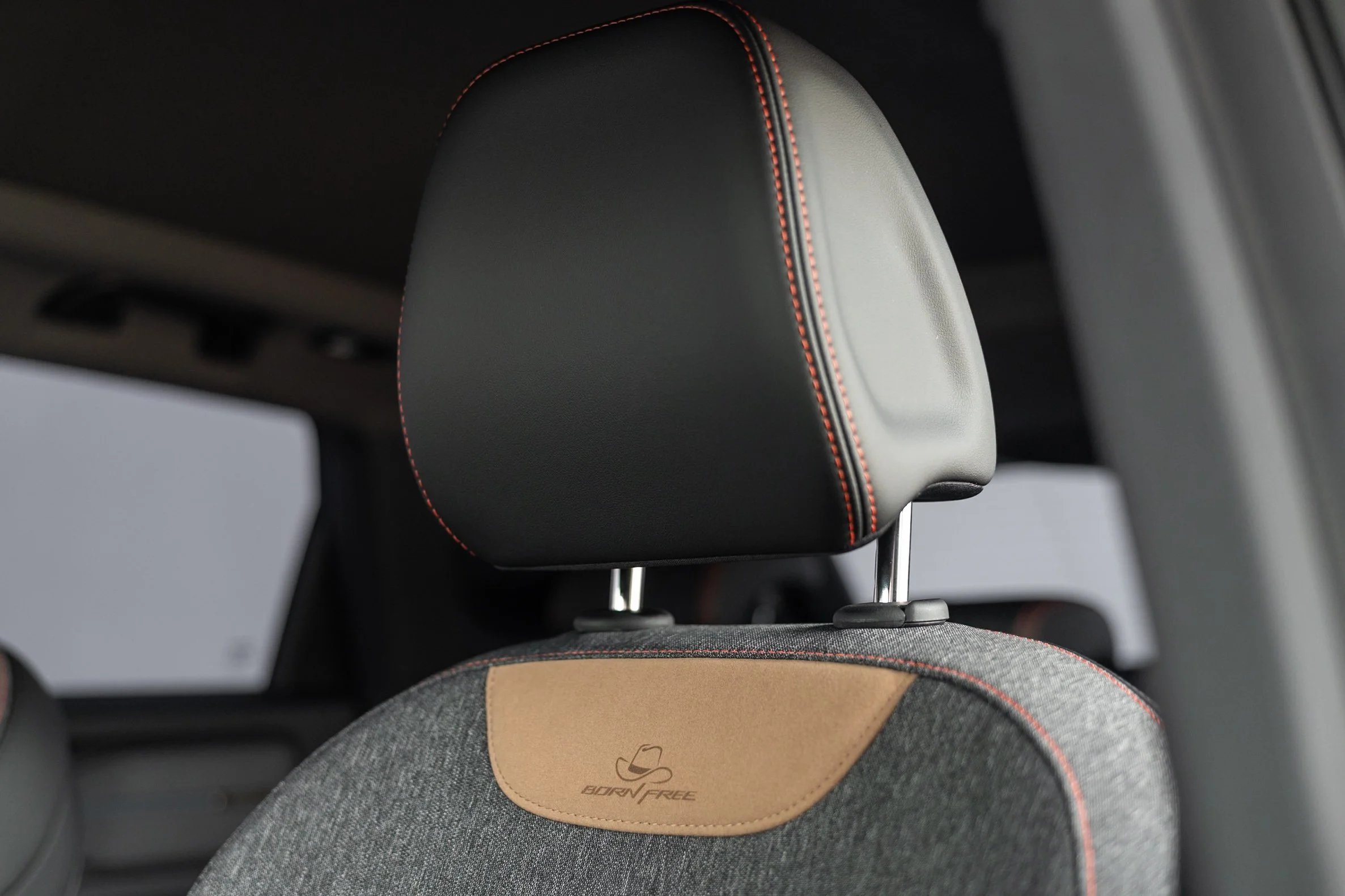 Close-up of a stylish black car headrest with red stitching, with a person's headrest and their head wearing a gray textured shirt and a beige leather travel pillow with a logo underneath.